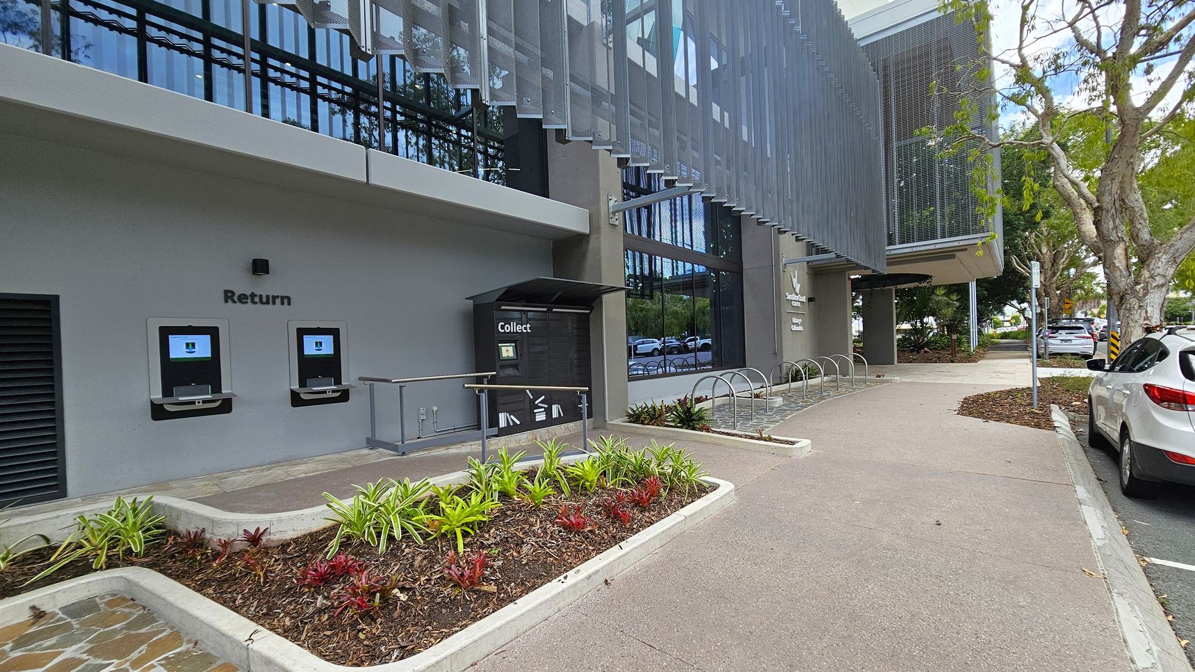 On the left, there are two automated return machines embedded in a grey wall, labelled "Return". Below them is a small garden bed with various plants. In the centre, a "Collect" locker is situated under a small awning. The building features large glass windows with a metal mesh awning above. On the right, a paved path leads past a bicycle rack and parked cars. Trees line the street, providing shade.