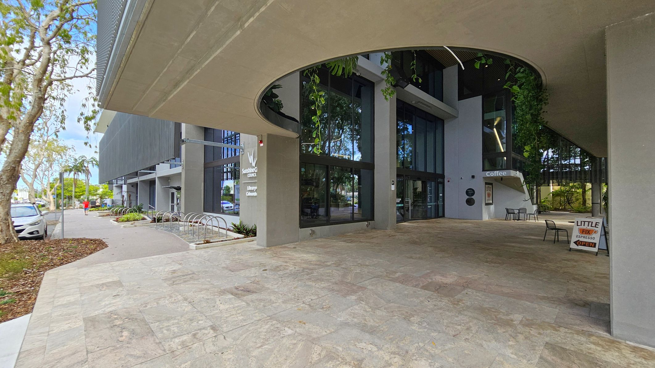 A modern building with large glass windows and a curved overhang. On the right, there's a sign for "Little Fix Espresso" and a few chairs and tables. The left side shows a pathway with bike racks and a parked car. Trees line the street, and a person in a red shirt walks along the pavement. The floor is tiled in a mix of green and brown shades.
