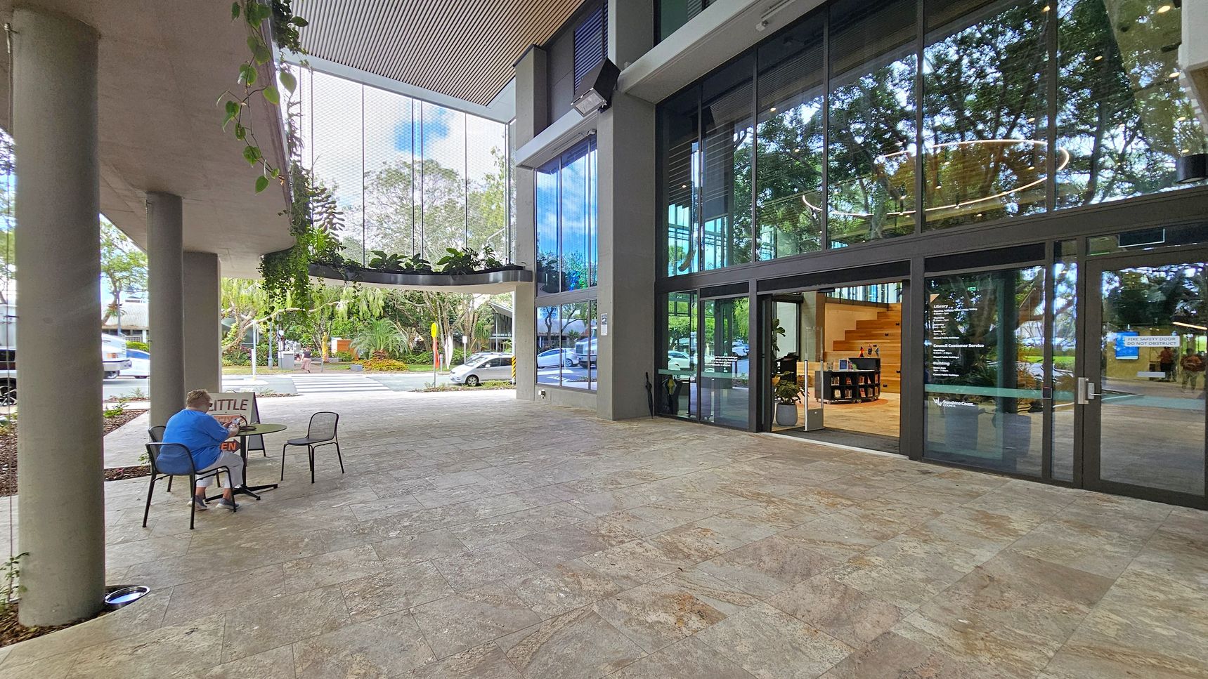 A modern outdoor area with a grey tiled floor. On the left, a person in a blue shirt sits at a small table with a sign that reads "LITTLE" and "OPEN". There are two empty chairs nearby. Above, hanging plants are suspended from the ceiling. On the right, large glass doors lead into a building with visible interior stairs and a small bookshelf. The background features a street with parked cars and lush greenery.