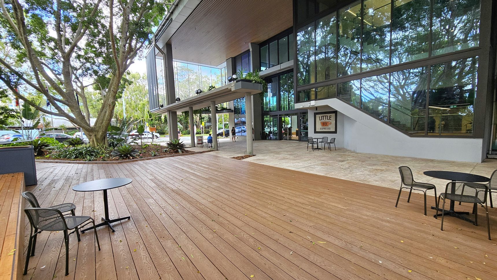 A spacious outdoor area with wooden decking on the left, featuring a round table and chairs. A large tree with a thick trunk and lush foliage is surrounded by plants. On the right, a modern building with large glass windows and a sign for "Little Fix Espresso" near the entrance. The building has a covered walkway supported by columns. The ground transitions from wood to stone tiles near the building.