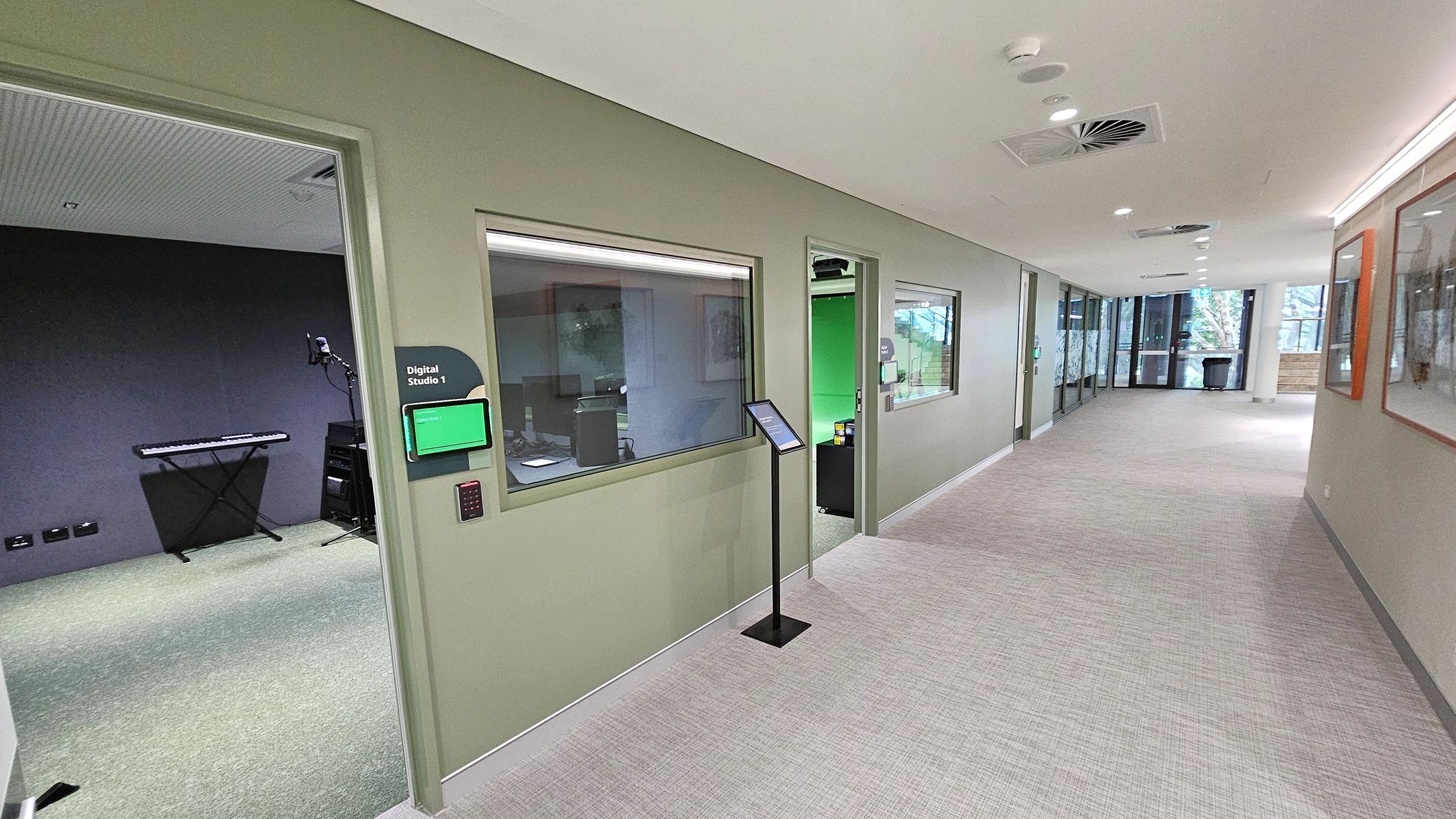 A corridor with light grey carpet and a light-coloured ceiling featuring recessed lighting and air vents. On the left, there are rooms with large windows and doors, one labelled "Digital Studio 1" with a digital display. Inside, a keyboard and microphone stand are visible. A stand with a tablet is positioned outside the room. On the right, framed pictures hang on the wall. At the end of the corridor, large windows reveal trees outside.