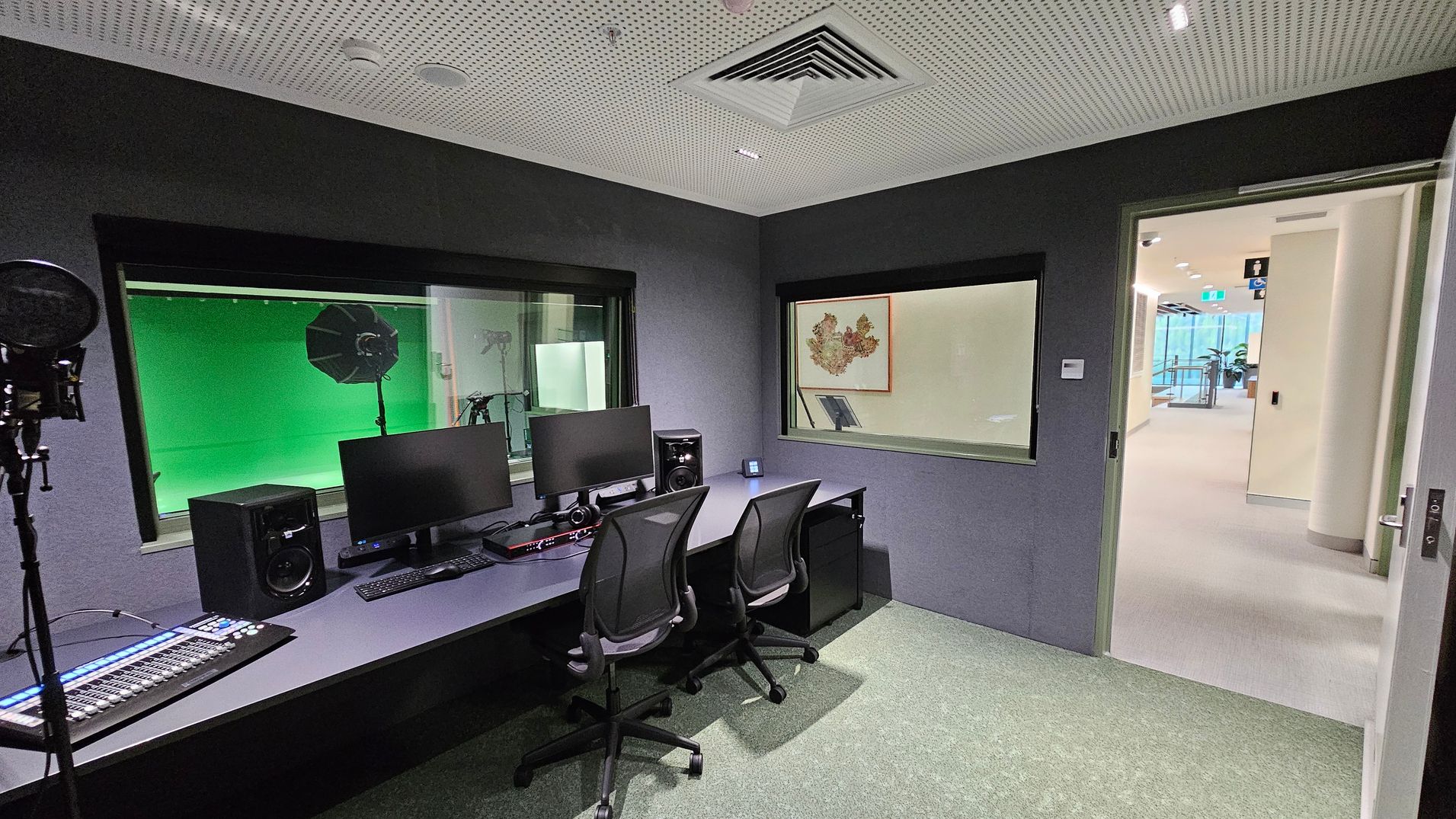 A recording studio with a dark interior. On the left, there's a microphone and a mixing console. In the centre, two black chairs face a desk with dual monitors, speakers, and headphones. A large window reveals a green screen and lighting equipment. On the right, a door opens to a bright hallway with visible plants and signage. The ceiling is perforated with ventilation, and the floor is carpeted.