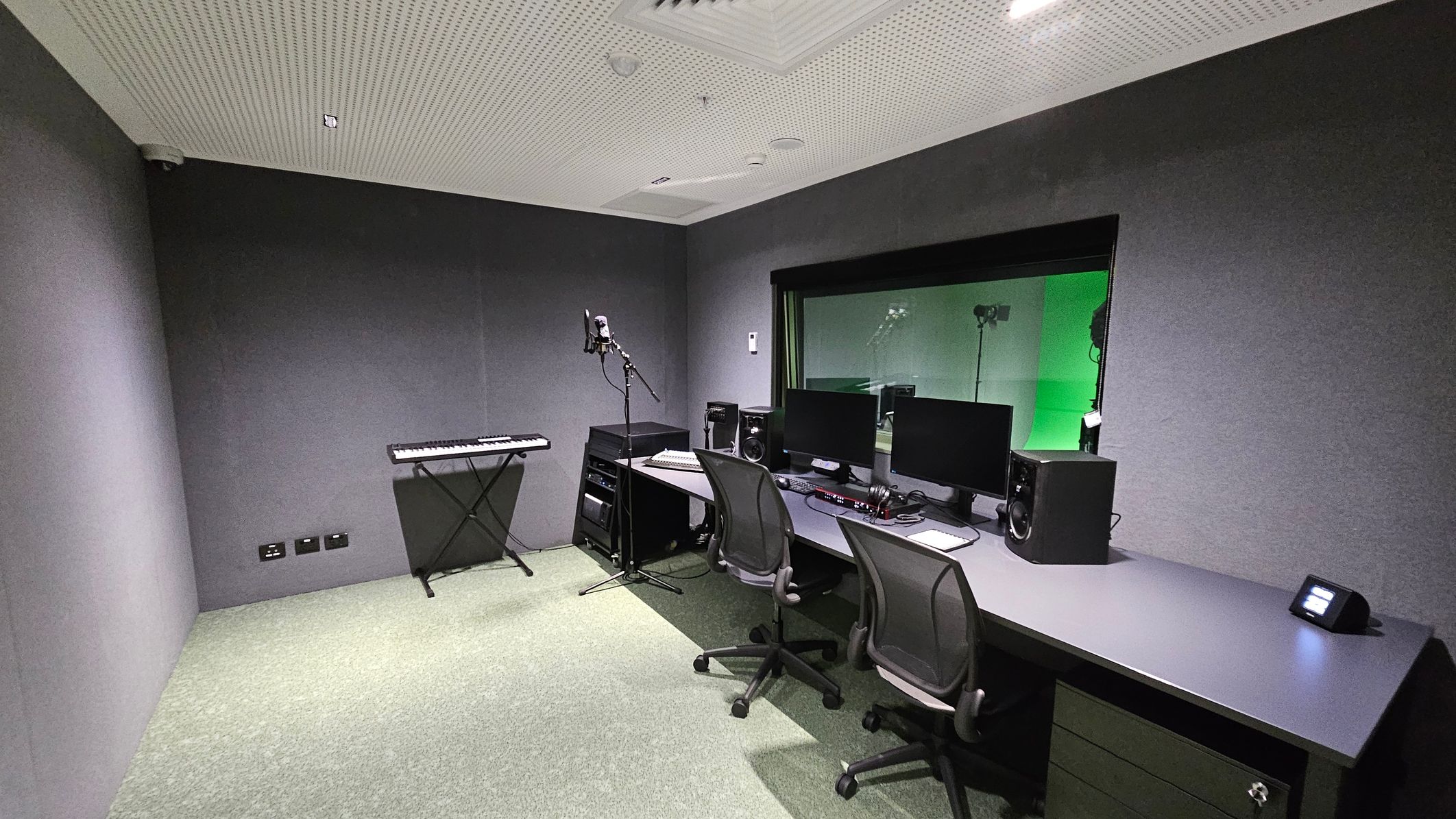 A recording studio with grey walls and a perforated ceiling. On the left, a keyboard on a stand and a microphone on a stand. In the centre, a rack with audio equipment. On the right, a long desk with two office chairs, two computer monitors, speakers, and headphones. A large window shows a green-lit room beyond. The floor is carpeted in a light grey-green colour.