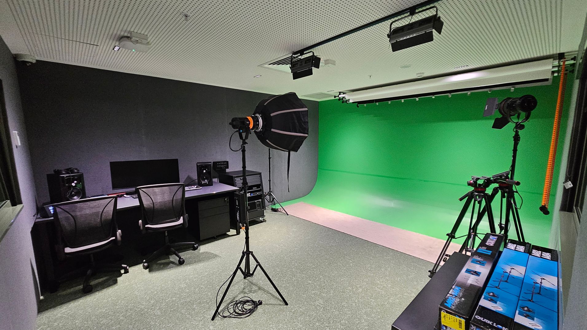 A room with a green screen backdrop on the right side, extending from the floor to the ceiling. In the centre, there's a camera on a tripod and a large studio light with a softbox. On the left, a desk with two chairs, a computer monitor, and speakers. The ceiling is perforated with mounted lights. The floor is carpeted in grey. In the foreground, there are boxes stacked on a table.