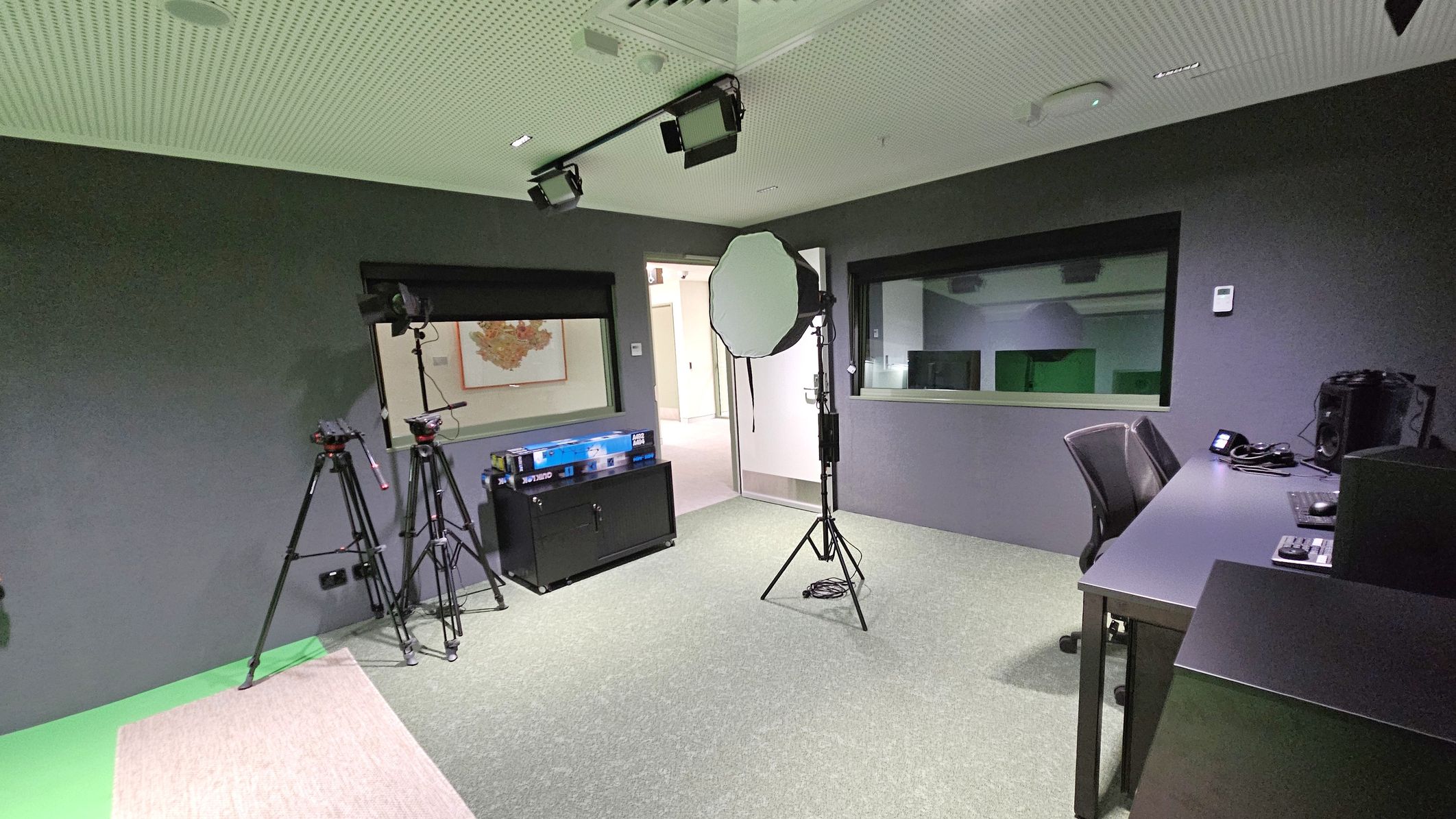 A room with a grey carpeted floor and a perforated ceiling. On the left, two tripods stand near a black cabinet with a box on top. In the centre, a large softbox light is positioned near an open door. On the right, a desk with a chair holds a computer, keyboard, and other equipment. A large window is set into the wall, providing a view into another room. The walls are dark grey, and there are ceiling-mounted lights.