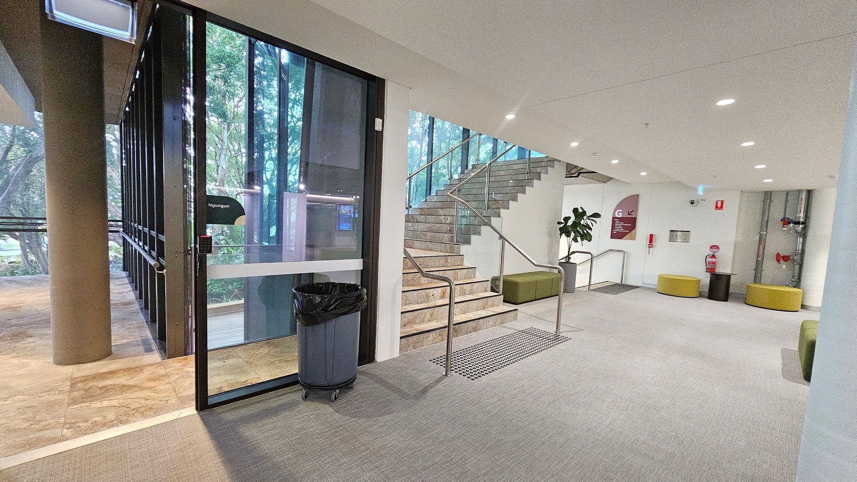 A modern indoor space with a glass door on the left, leading outside. A cylindrical column is visible near the door. In the centre, a staircase with a metal railing ascends, flanked by a green cushioned bench. The right side features a white wall with a fire extinguisher, yellow circular seats, and exposed pipes. The floor is carpeted, and the ceiling has recessed lighting. A potted plant is near the stairs.