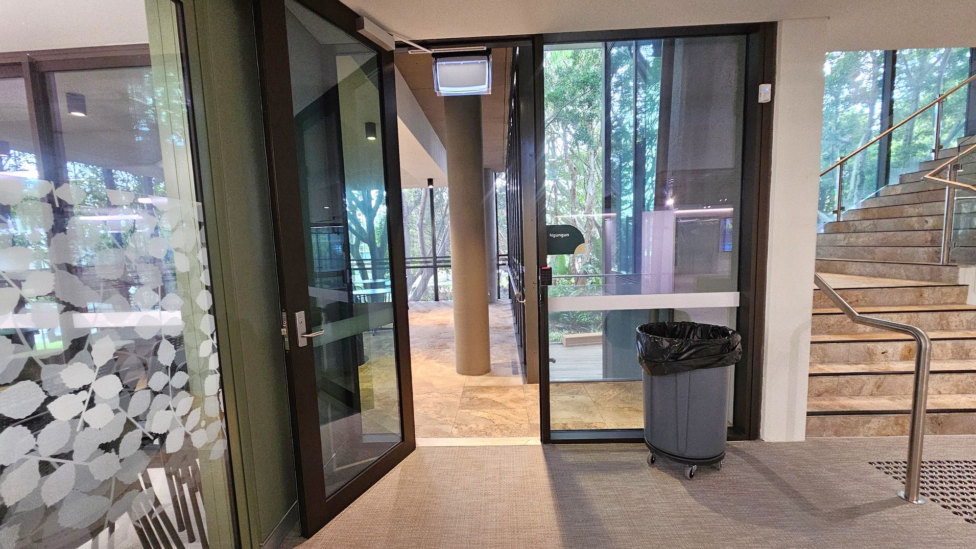 Glass doors with a leaf pattern on the left, leading to an outdoor area with trees visible through the glass. A grey rubbish bin with a black liner is positioned near the door. To the right, a staircase with a metal handrail ascends. The floor is carpeted, and the ceiling is plain.