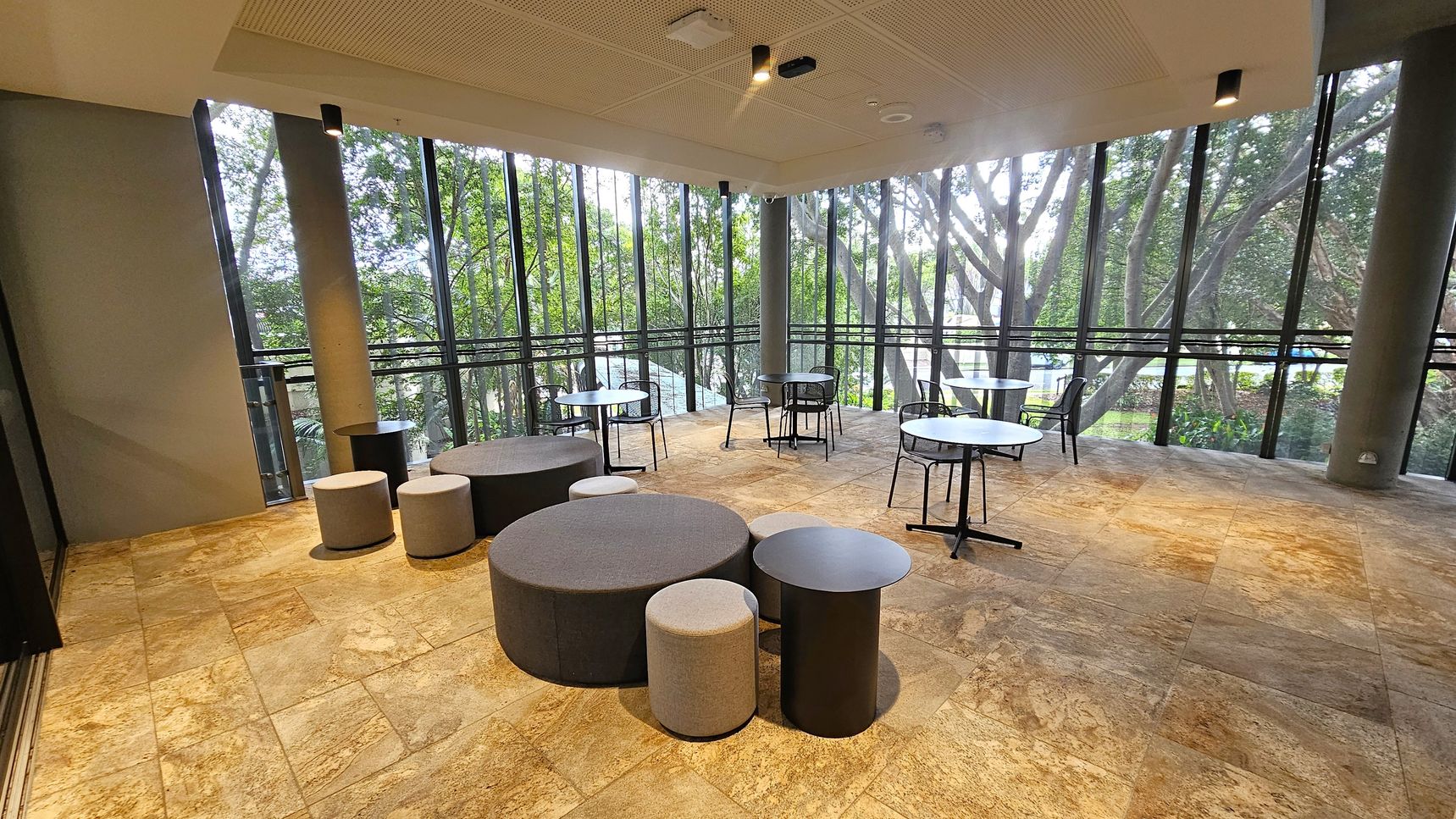 A room with large floor-to-ceiling windows showcasing a view of lush green trees outside. The floor is covered with light brown tiles. In the centre, there are round tables with black bases and matching chairs. To the left, there are circular cushioned stools and a low round table. The ceiling is white with recessed lighting.