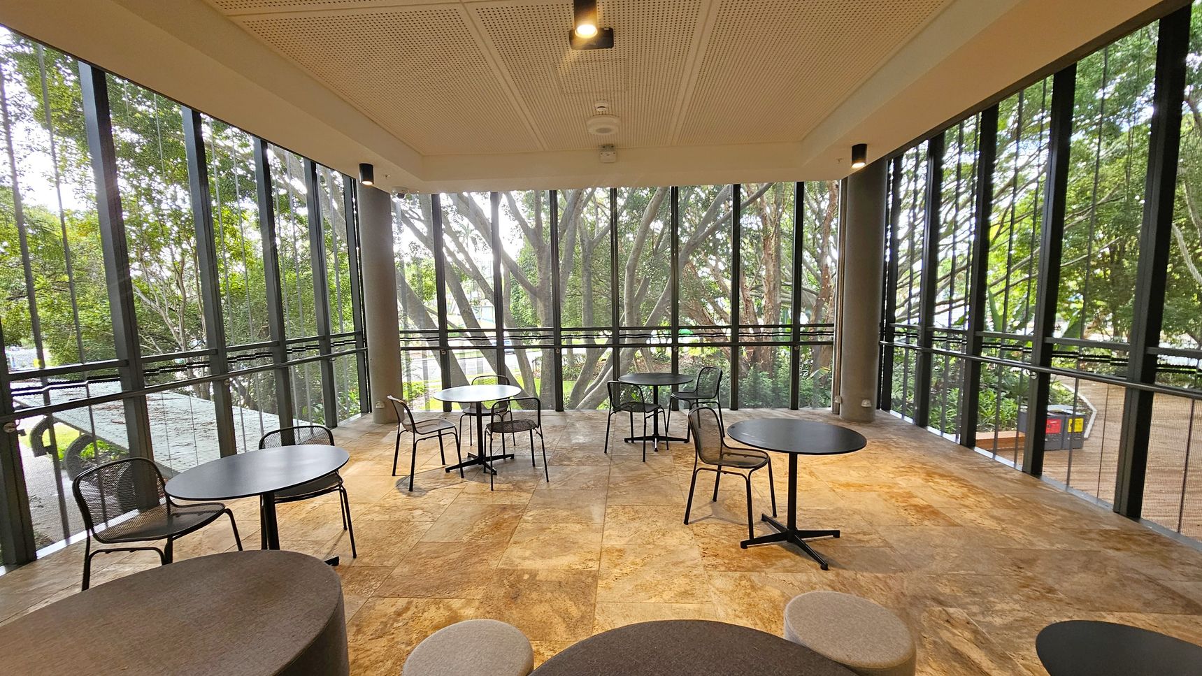 A spacious indoor area with large floor-to-ceiling windows on three sides, offering a view of lush green trees outside. The floor is covered with stone tiles in earthy tones. Several round tables with black metal chairs are scattered throughout the space. The ceiling is white with recessed lighting. The area is well-lit, with natural light streaming in through the windows.