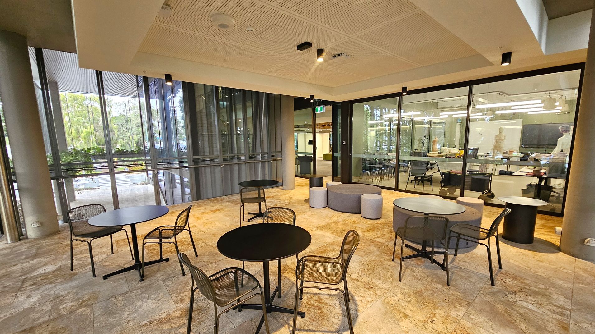A modern indoor seating area with round black tables and mesh chairs on a stone-patterned floor. On the right, there are grey and purple cushioned stools. The background features large glass windows and doors, revealing a well-lit room with tables, chairs, and a whiteboard. The ceiling is light-coloured with recessed lighting and ventilation fixtures. Outside the windows, greenery is visible.