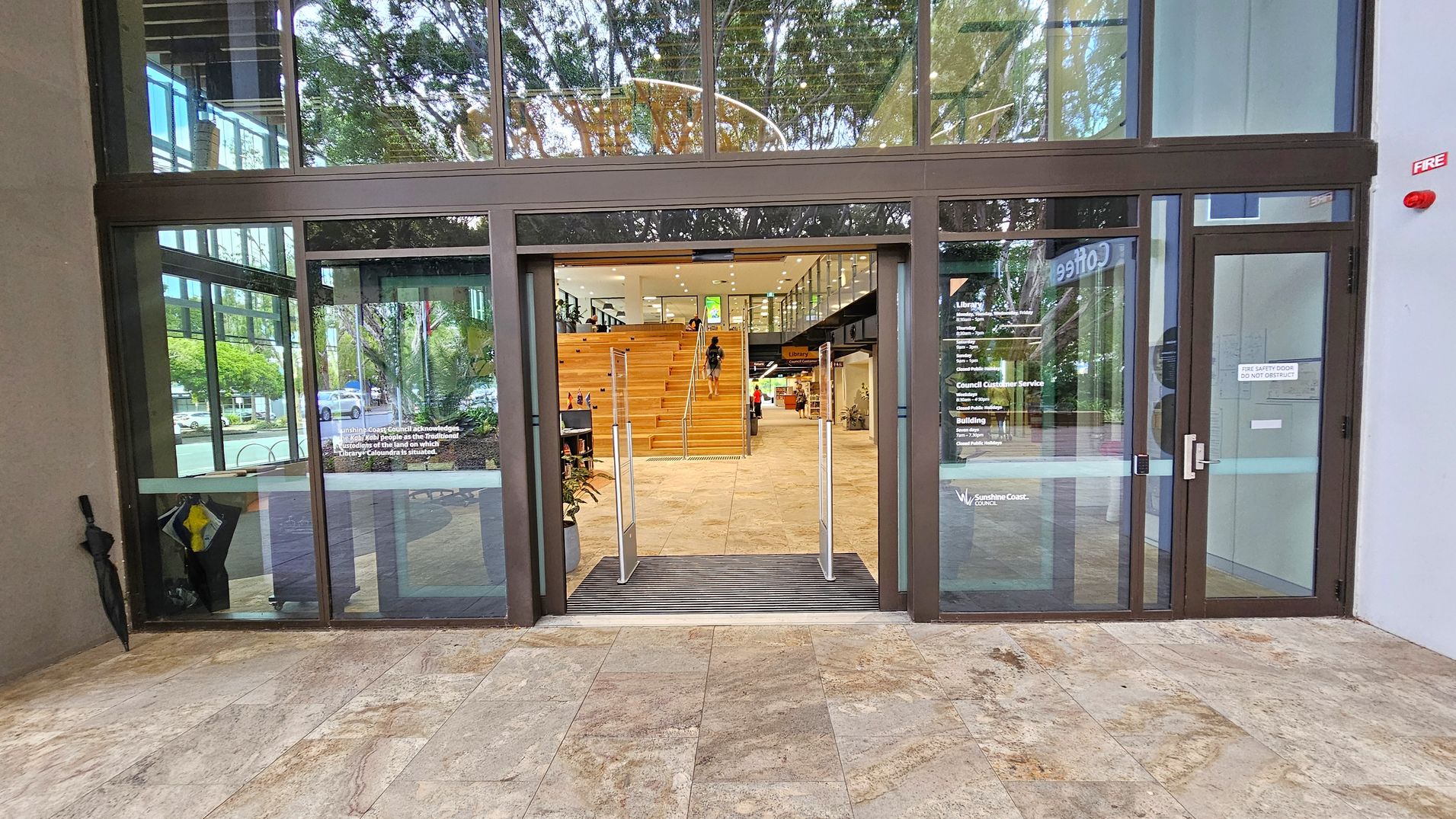 Glass entrance doors with a metal frame lead into a building. Inside, a wide staircase with wooden steps is visible, with a person ascending. To the right, there is a sign indicating a library and other facilities. The floor outside is tiled with a stone pattern. Reflections of trees and the street are visible on the glass. An umbrella is propped against the wall on the left, and a fire safety sign is on the right door.