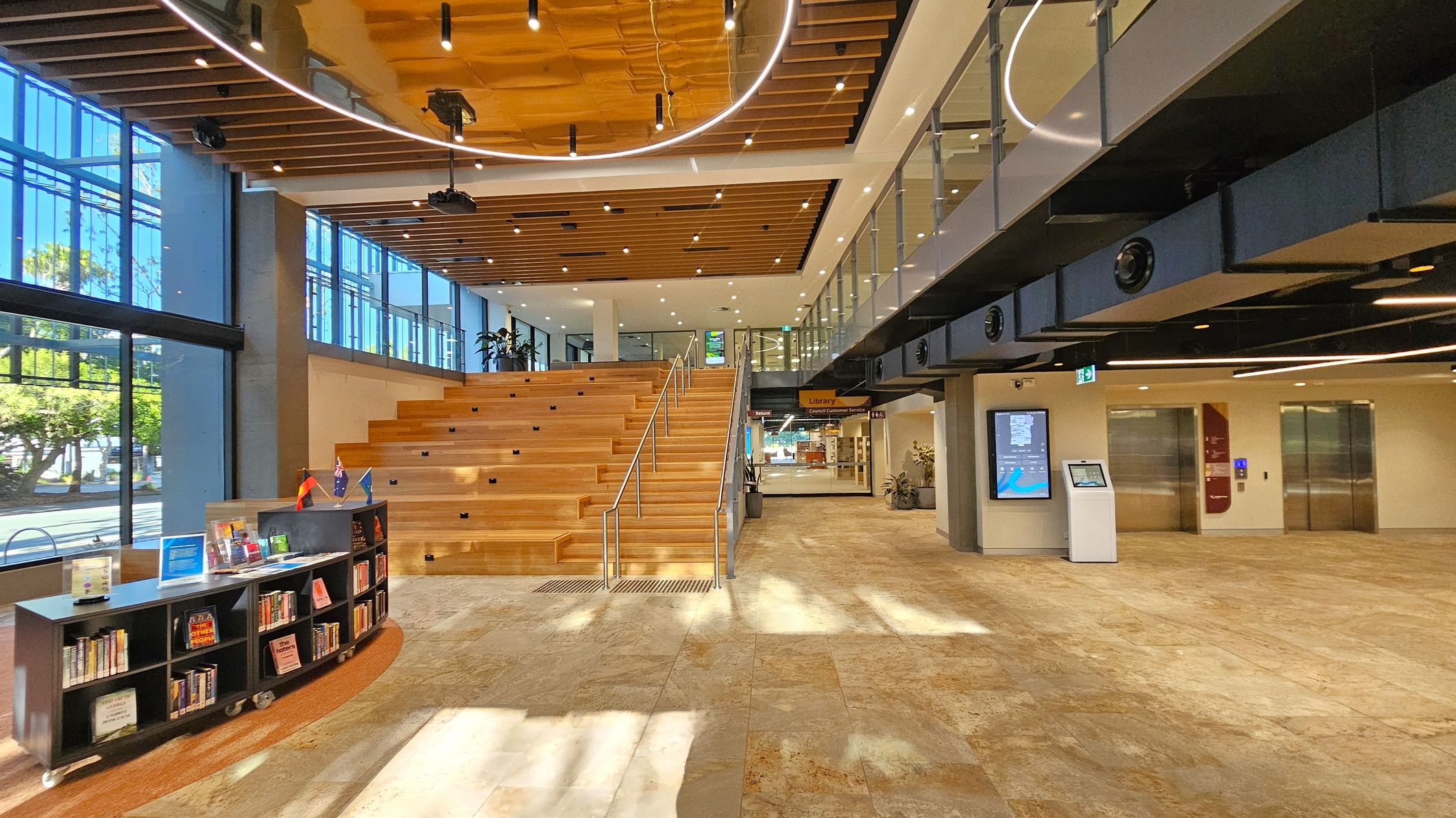 A spacious, modern interior with a large wooden staircase in the centre, leading to an upper level with glass railings. On the left, there is a bookshelf with various books and small flags on top. Large windows allow natural light to flood in, illuminating the stone-tiled floor. On the right, there are two elevators and a digital information kiosk. The ceiling features wooden slats and circular lighting fixtures.