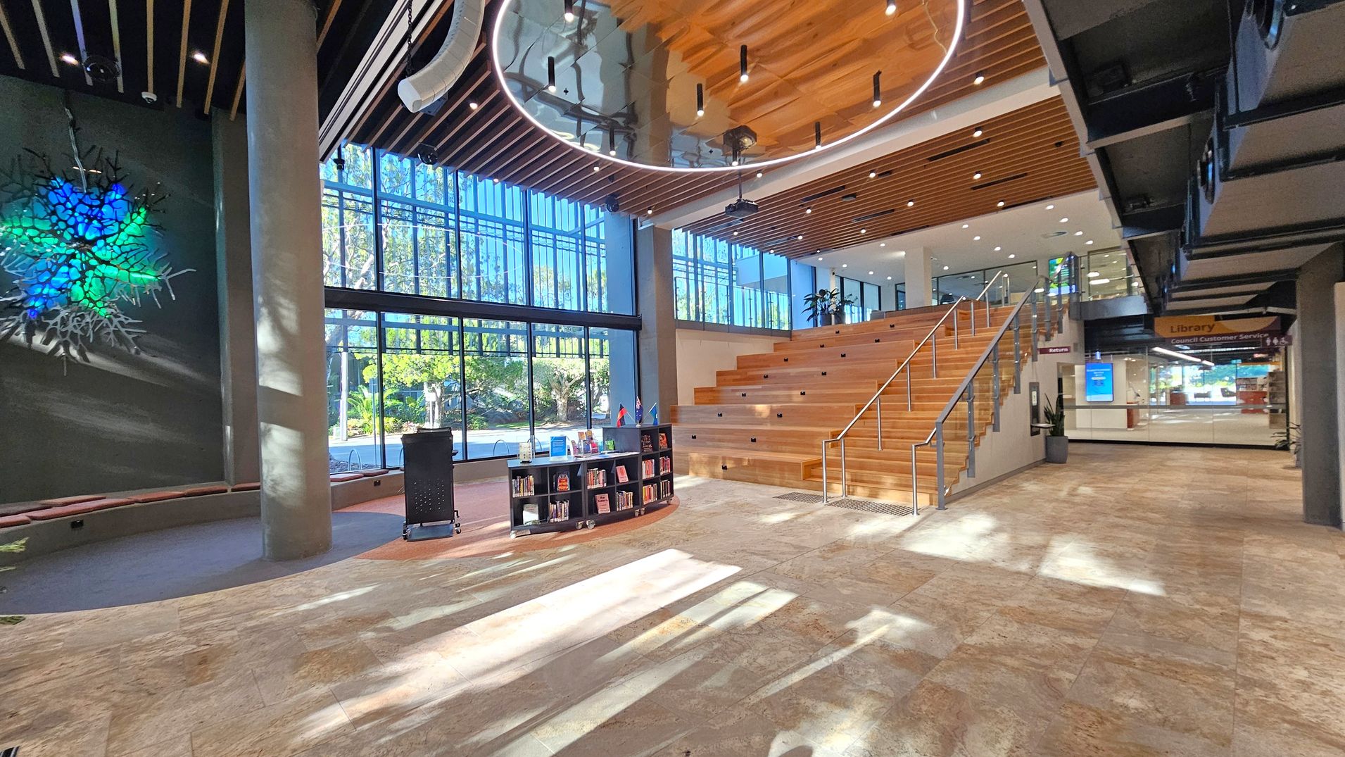 A spacious interior with a modern design. On the left, a large wall features an illuminated abstract art piece in blue and green. A tall column stands nearby. In the centre, a small bookshelf is positioned on a light-coloured tiled floor. To the right, a wide wooden staircase with metal railings leads to an upper level. The background has large glass windows allowing natural light to flood in. The ceiling has wooden slats and circular lighting fixtures.