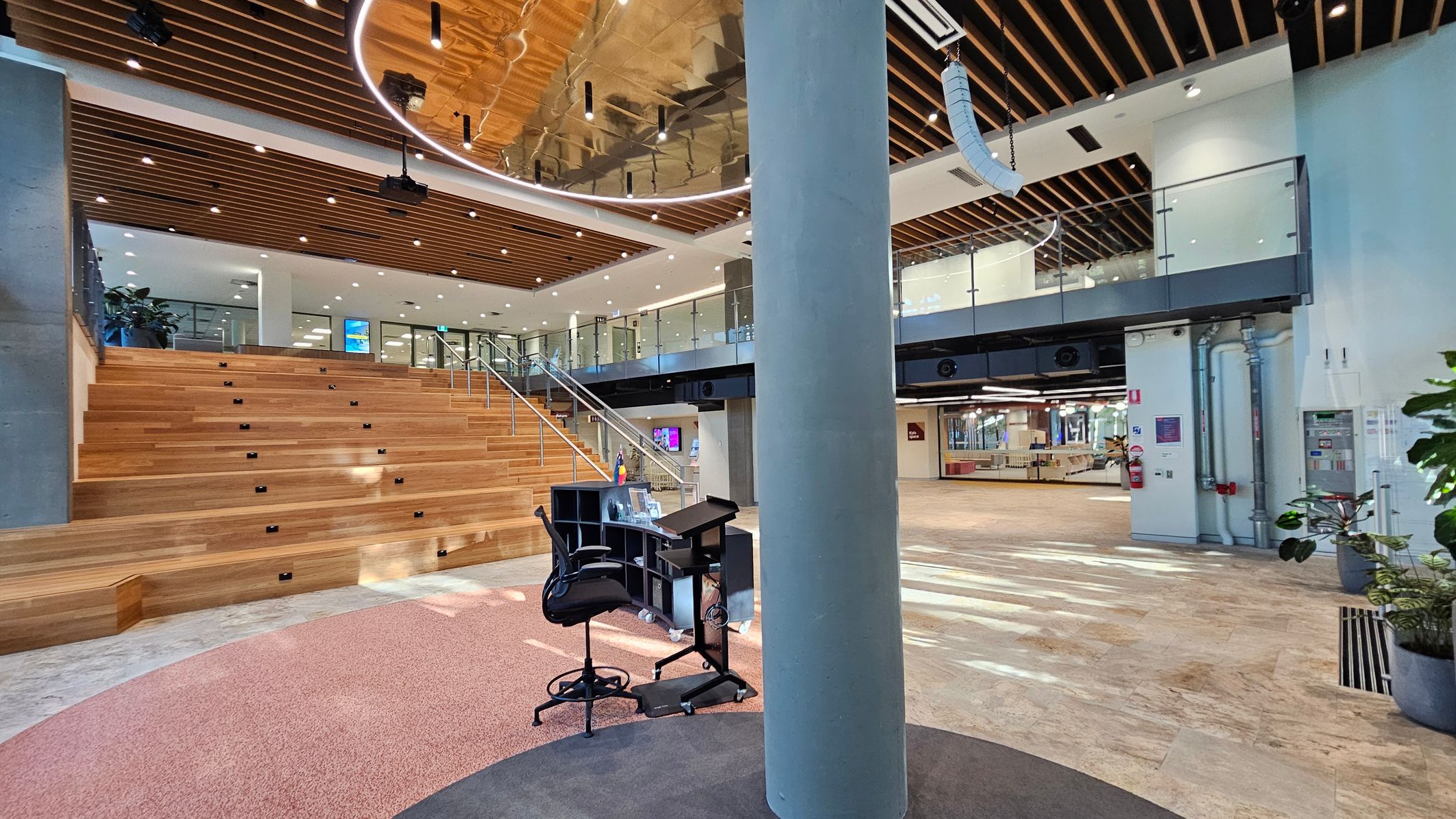 A modern interior space with a wooden staircase on the left leading to a mezzanine level with glass railings. The ceiling features wooden slats and recessed lighting. In the centre, a large cylindrical column stands near a small desk and chair on a circular carpet. The floor is a mix of light-coloured tiles and carpet. On the right, there are plants and visible pipes, with open space leading to other areas.