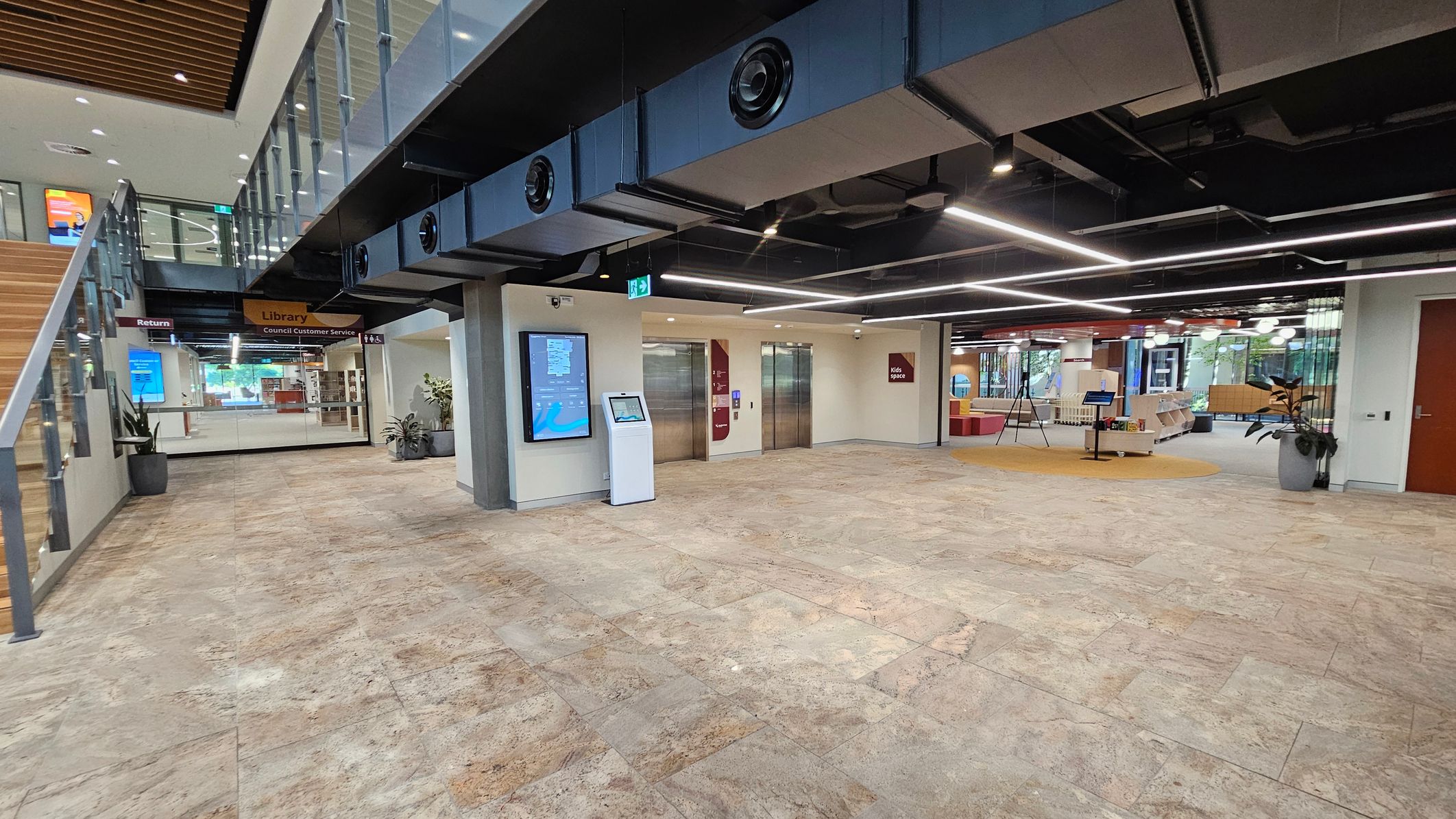 A modern interior space with a stone-tiled floor. On the left, a staircase with glass railings leads to an upper level. Below the stairs, a sign indicates the library and council customer service area. In the centre, two lifts are flanked by digital screens and a kiosk. On the right, a seating area with bookshelves and a circular carpet is visible. The ceiling features exposed ductwork and linear lighting. Potted plants are placed near the walls.