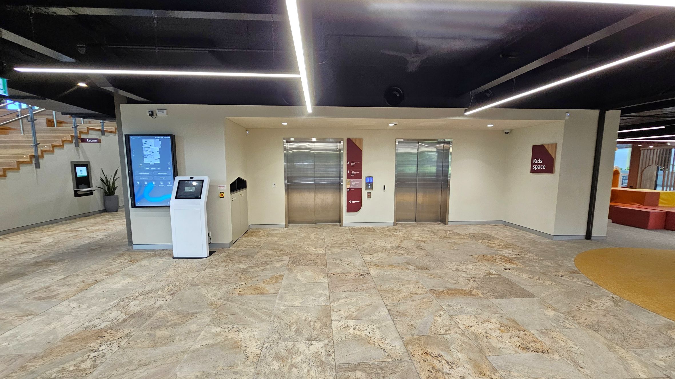 Two lifts with metallic doors are centred on a light-coloured wall. To the left, a digital directory and a white kiosk stand near a staircase with wooden steps and a potted plant. On the right, a sign reads "Kids space" above a colourful seating area. The floor is tiled with a mix of beige and green hues, and the ceiling features black panels with linear lighting.