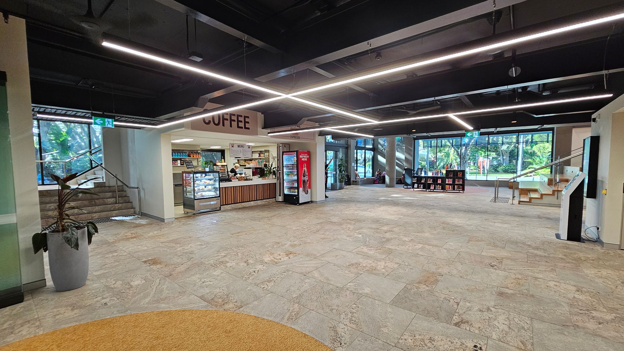 A spacious indoor area with a coffee shop on the left, featuring a counter with a display case and a drinks fridge. Above, a sign reads "COFFEE". The floor is tiled in a light stone pattern. In the background, large windows reveal greenery outside. On the right, there are stairs leading up, and a small bookshelf is visible. The ceiling is dark with linear lights. A potted plant is on the left near the stairs.