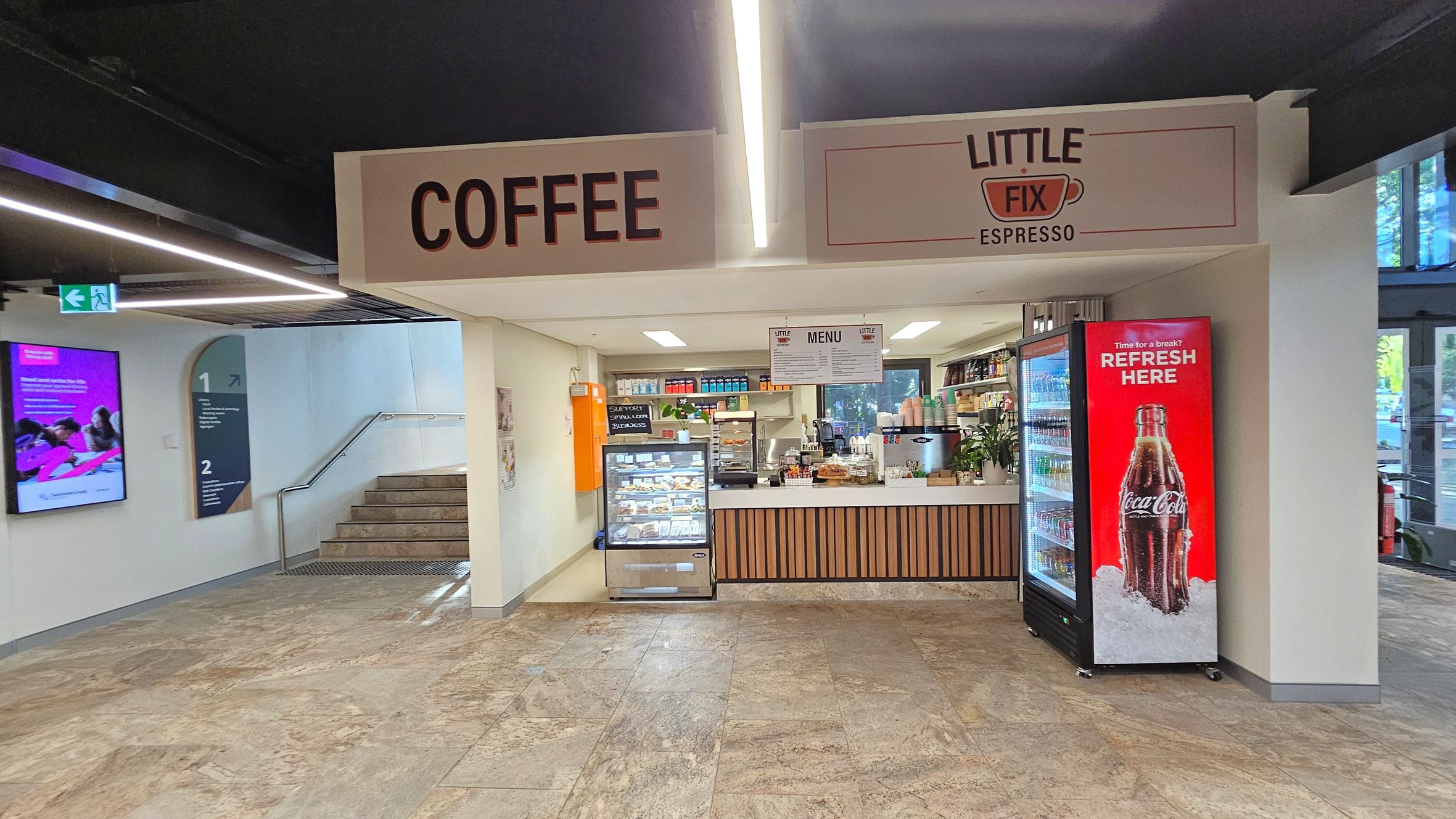 A small coffee shop with a sign reading "COFFEE" and "LITTLE FIX ESPRESSO" above. On the right, a red Coca-Cola branded refrigerator displays drinks. In the centre, a counter with a menu above offers various items, and a glass display case shows pastries. Shelves with products are visible in the background. On the left, a staircase leads upwards, and a digital screen displays information. The floor is tiled, and the ceiling has recessed lighting.