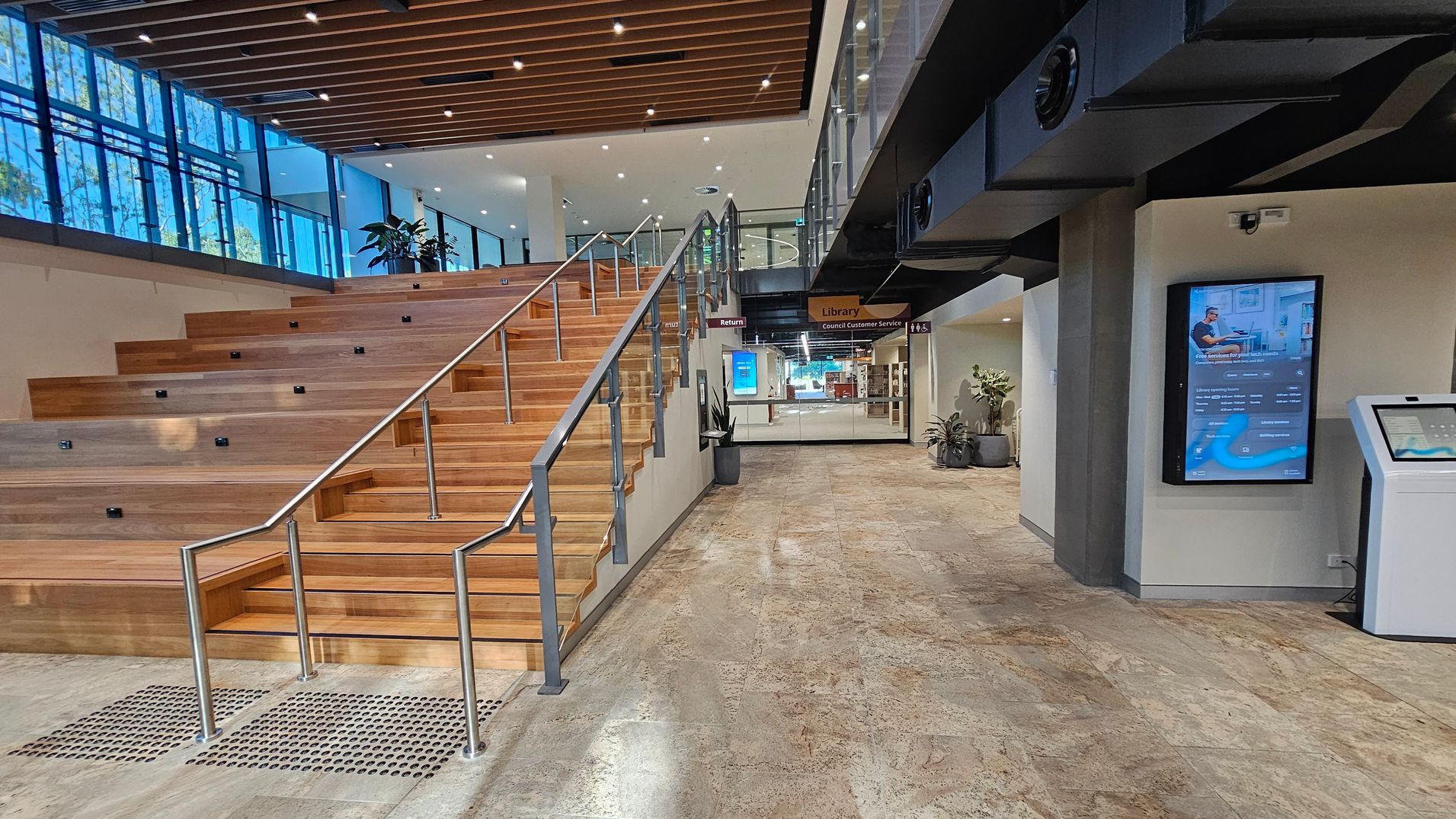 A modern interior space with a wide wooden staircase on the left, featuring metal railings. Large windows with blue-tinted glass line the upper left wall. On the right, a digital information screen is mounted on the wall above a kiosk. The floor is tiled in a light stone pattern. In the background, signs indicate directions to a library and customer service area, with potted plants adding greenery. The ceiling has wooden slats with recessed lighting.