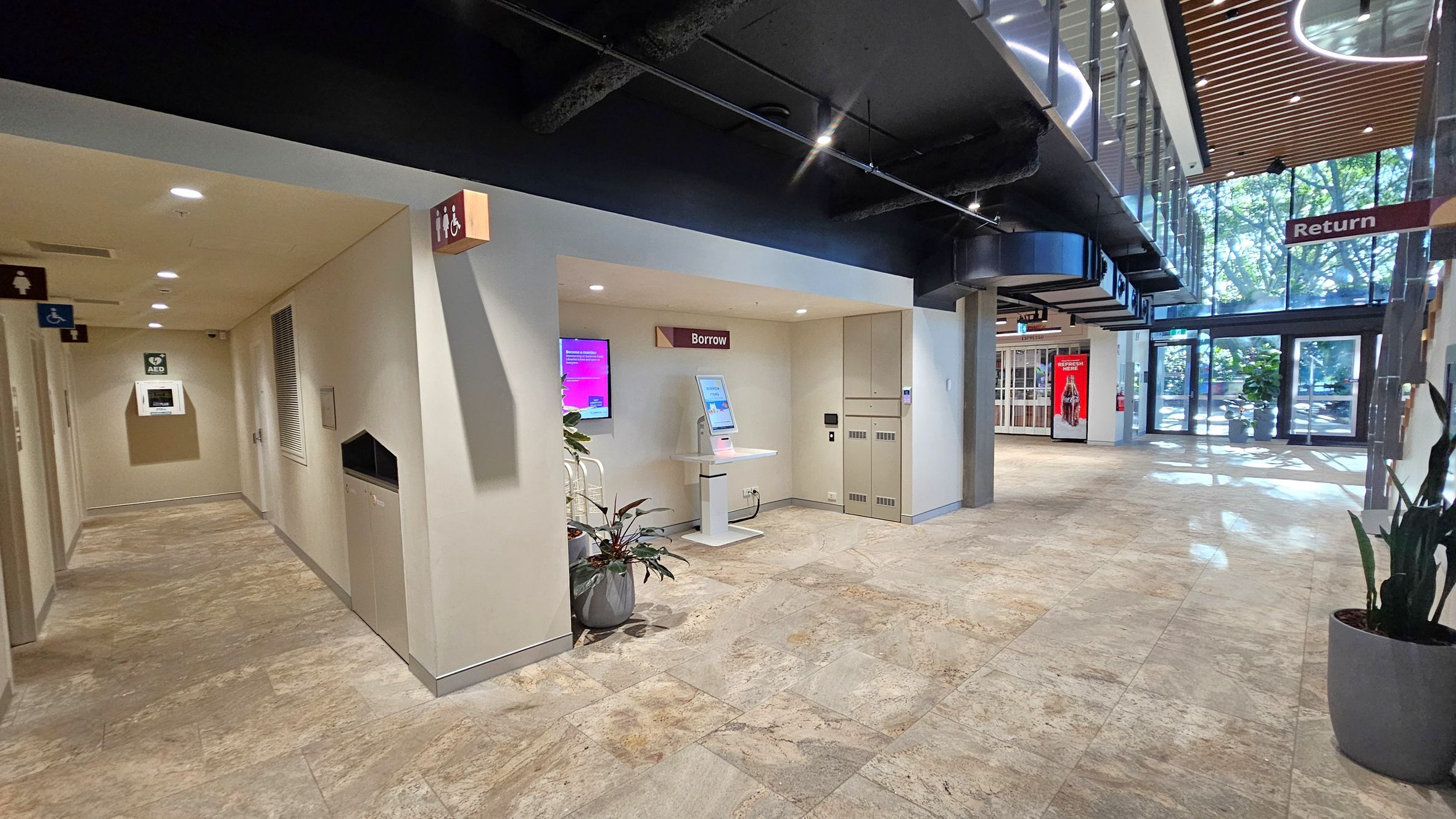 A modern interior space with a light-coloured tiled floor. On the left, a hallway leads to restrooms, marked by signs, and features an AED on the wall. In the centre, a kiosk labelled "Borrow" is positioned against a wall with a digital screen and a potted plant nearby. On the right, large glass doors allow natural light to flood in, with a "Return" sign hanging above. The ceiling has exposed black ductwork and wooden slats.