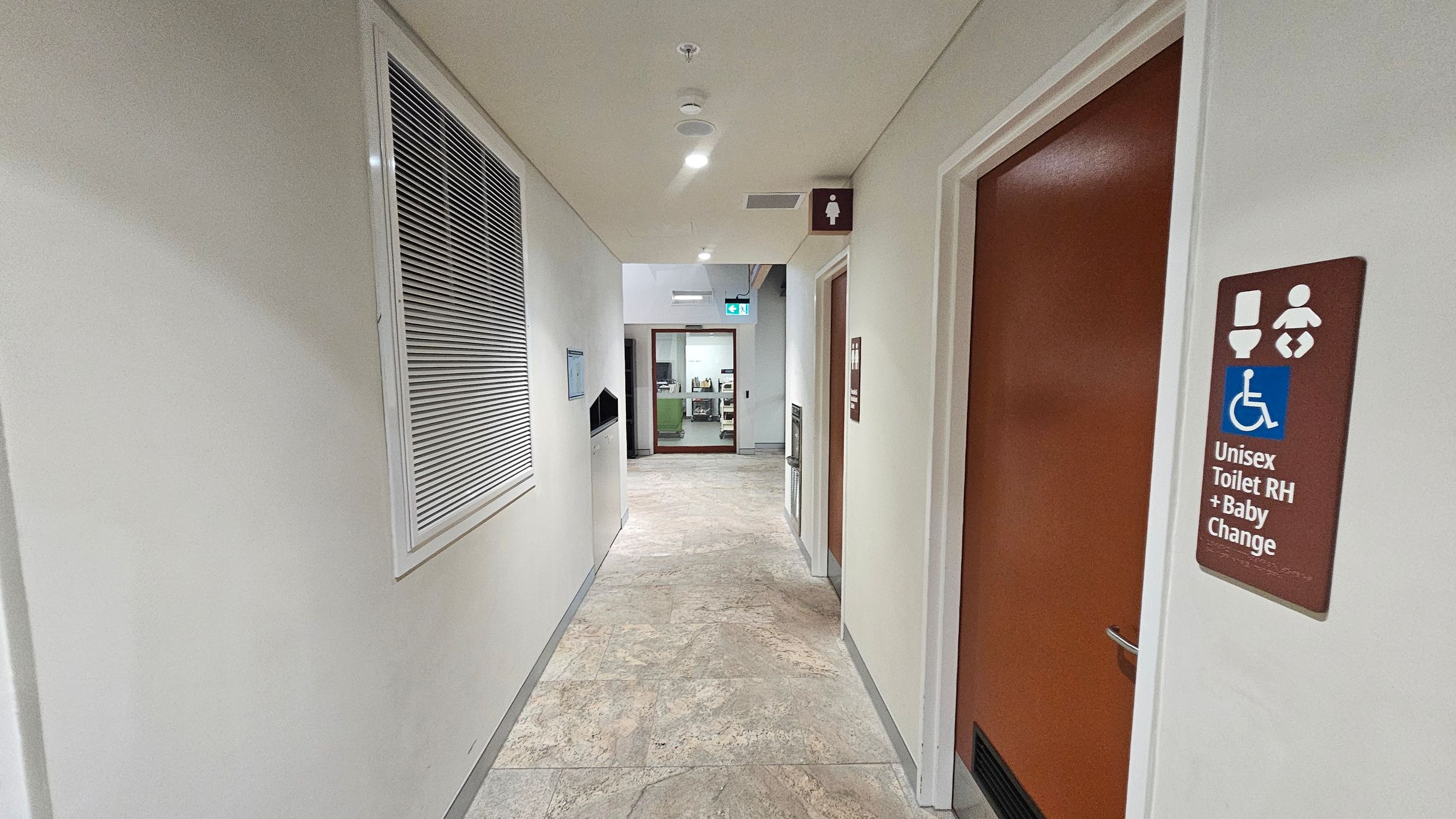 A corridor with a light-coloured ceiling and patterned floor tiles. On the right, there is a brown door with a sign indicating a unisex toilet with baby changing facilities and accessibility features. On the left, a wall vent and a waste disposal unit are visible. At the end of the corridor, a glass door leads to another room, with an illuminated exit sign above it.