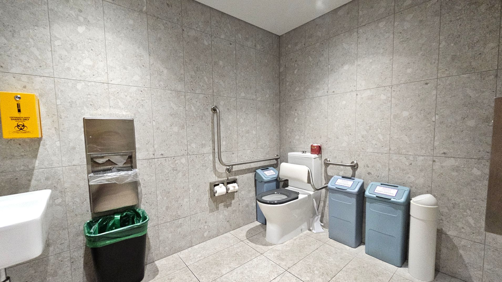 A public restroom with grey tiled walls and floor. On the left, there's a white sink and a yellow sharps disposal box mounted on the wall. Below it, a paper towel dispenser is above a black bin with a green liner. In the centre, a white toilet with a black seat is equipped with a support rail. To the right of the toilet, there are two blue sanitary bins and a white bin. The ceiling is plain and white.