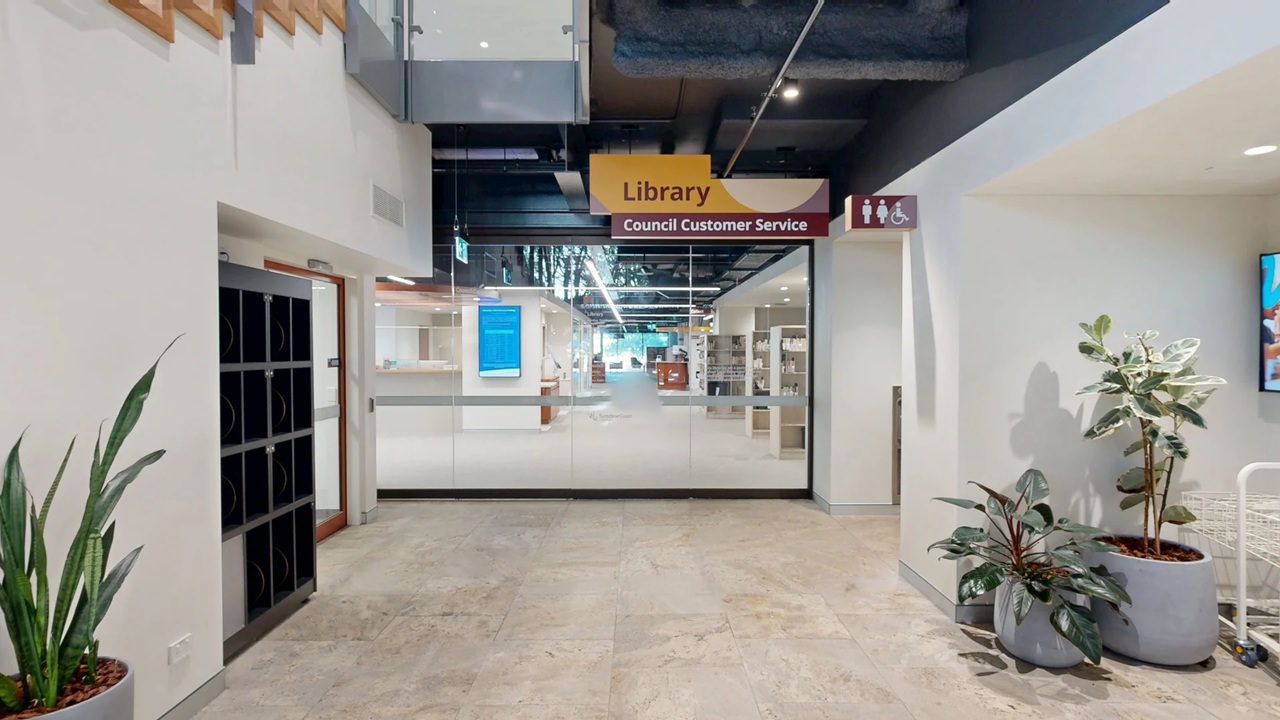 Entrance to a library with a glass door in the centre, marked by a sign reading "Library Council Customer Service". On the left, there are lockers and a tall potted plant. On the right, two potted plants are placed near a wall with a digital screen. The floor is light-coloured, and the ceiling is dark with exposed fixtures. Signs for restrooms are visible above the entrance.