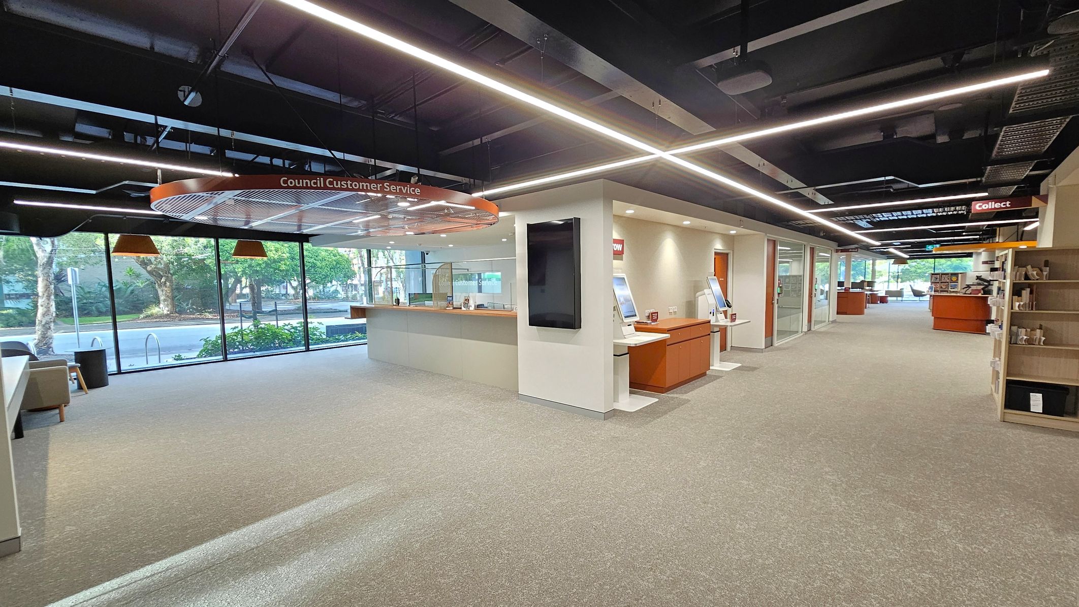 A modern interior space with a council customer service area on the left, featuring a counter and a sign above. Large windows provide a view of trees outside. On the right, there are self-service kiosks and a shelving unit. The ceiling has exposed beams and linear lighting. The floor is carpeted in a neutral tone, and the space is well-lit with a clean, organised layout.