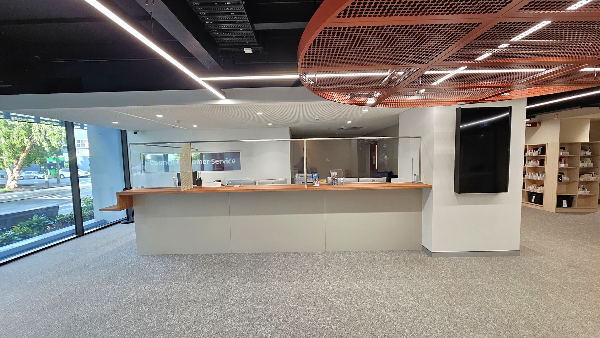A modern customer service area with a long counter in the centre, featuring a wooden top and glass partitions. Behind the counter are office chairs and a sign reading "Customer Service". On the right, a large black screen is mounted on a light green wall. Shelving with various items is visible further to the right. The left side has large windows with a view of trees and parked cars outside. The ceiling has a grid-like design with integrated lighting, and the floor is carpeted in grey.