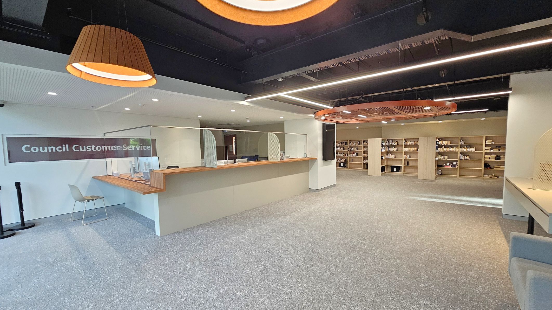A modern interior with a "Council Customer Service" desk on the left, featuring a wooden counter and a chair. Above, a large, circular wooden light fixture hangs from the ceiling. The right side has open shelving filled with books and items. The floor is carpeted in grey, and the ceiling is black with linear lighting. The space is open and well-lit, with a clean and organised appearance.