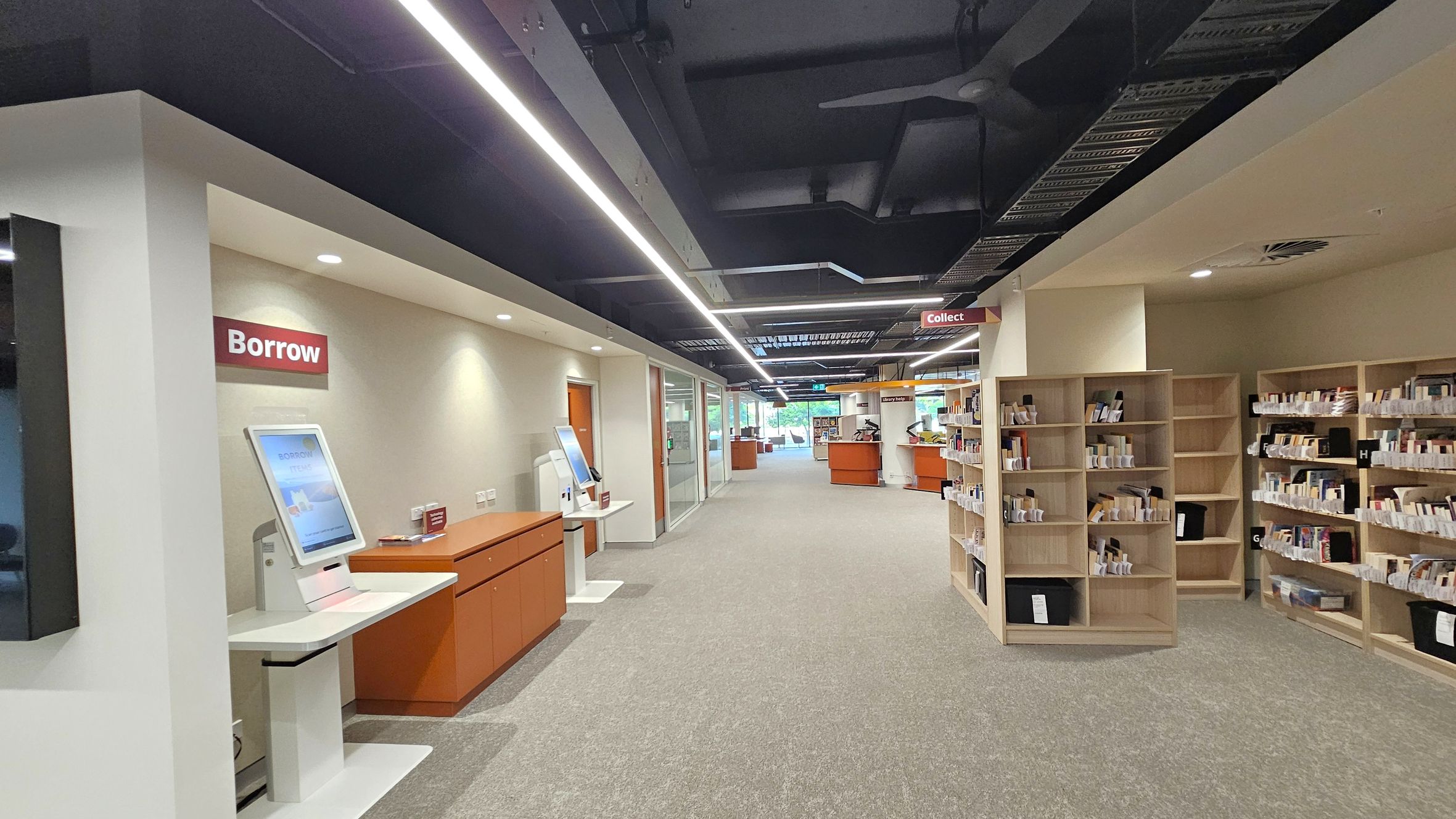 A library interior with a modern design. On the left, there's a "Borrow" station with a touchscreen kiosk and a counter. The floor is carpeted in grey. On the right, there are wooden bookshelves filled with books. In the middle, the space is open, leading to more shelves and a "Collect" area further back. The ceiling is dark with exposed ductwork and linear lighting.