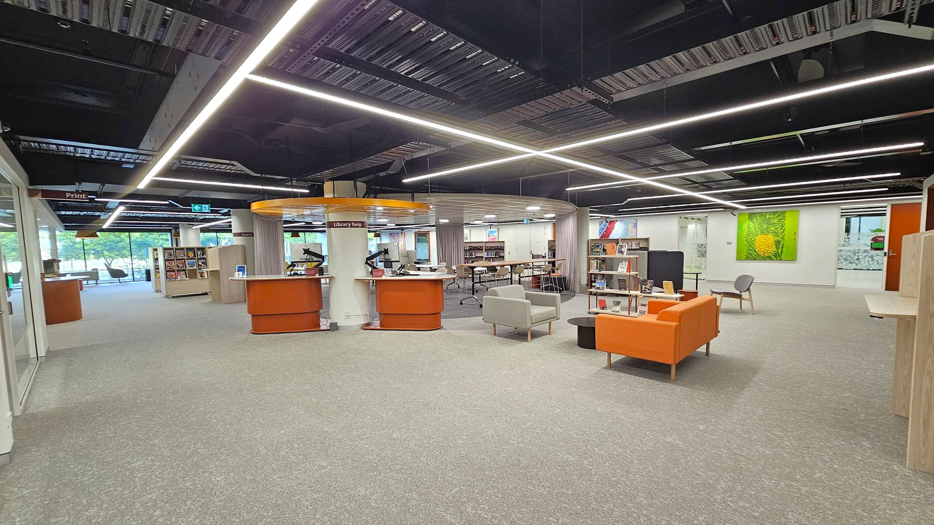 A modern library interior with a grey carpeted floor and an open ceiling featuring exposed ductwork and linear lighting. In the centre, there is a circular library help desk with orange accents. To the right, there are orange and grey seating areas with bookshelves. On the left, large windows provide natural light, and there are more bookshelves and seating. Bright artwork is displayed on the walls, adding colour to the space.