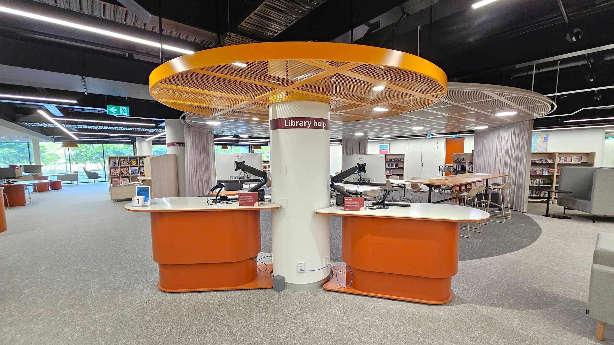 A modern library interior with a central "Library help" desk featuring two computer stations on orange counters. Above is a circular yellow structure with lighting. To the left, there are bookshelves and seating areas near large windows. On the right, a long table with chairs and more bookshelves. The floor is carpeted in grey and green, and the ceiling has exposed black beams with strip lighting.