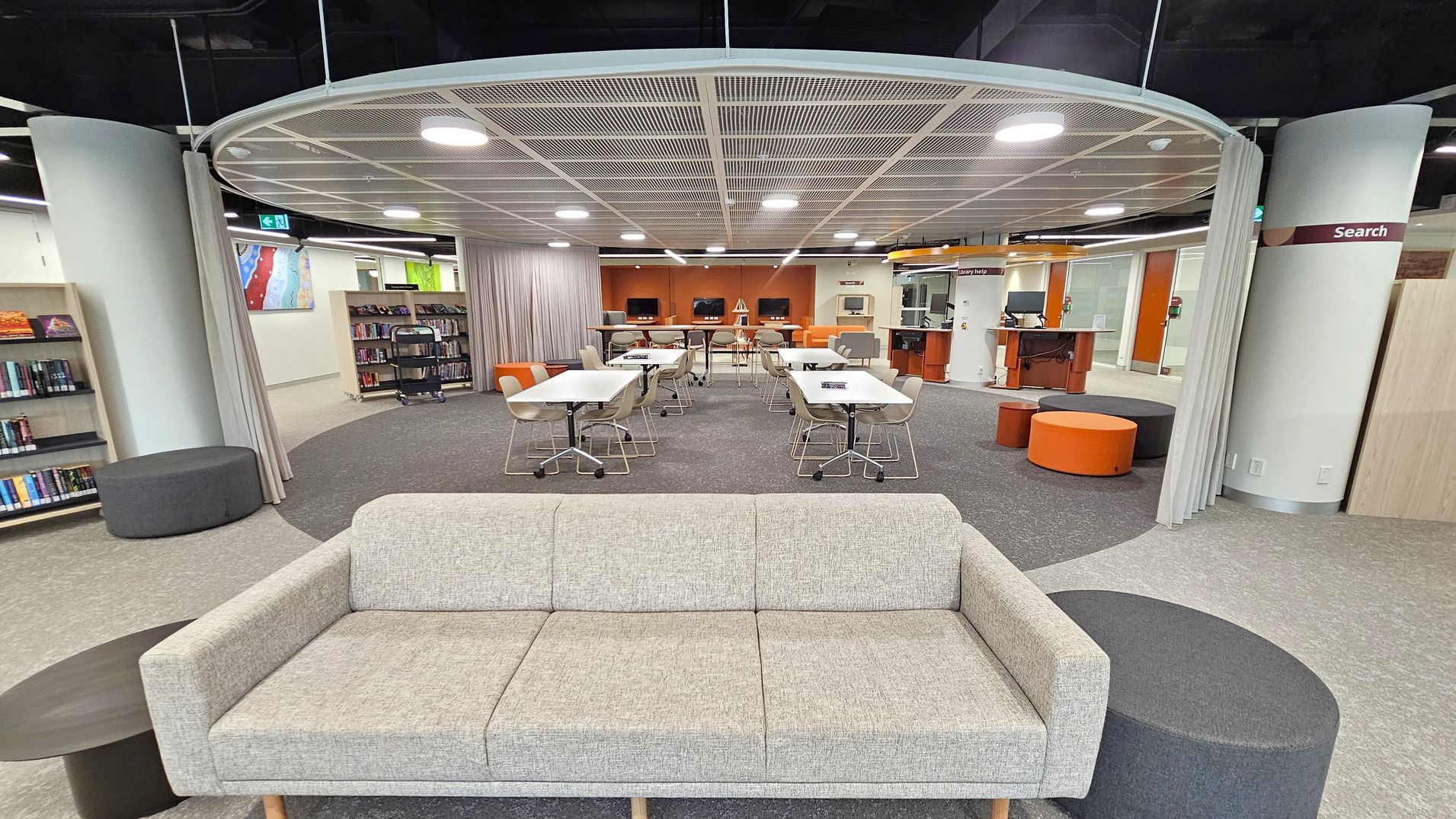 A modern library interior with a grey sofa in the foreground, accompanied by a round black side table. In the middle, there are several tables with chairs on wheels, and a few computer stations against the back wall. The ceiling features a grid pattern with circular lights. Bookshelves line the left side, and there are orange and black round stools scattered around. A sign on the right reads "Search". The floor is carpeted in grey and purple tones.