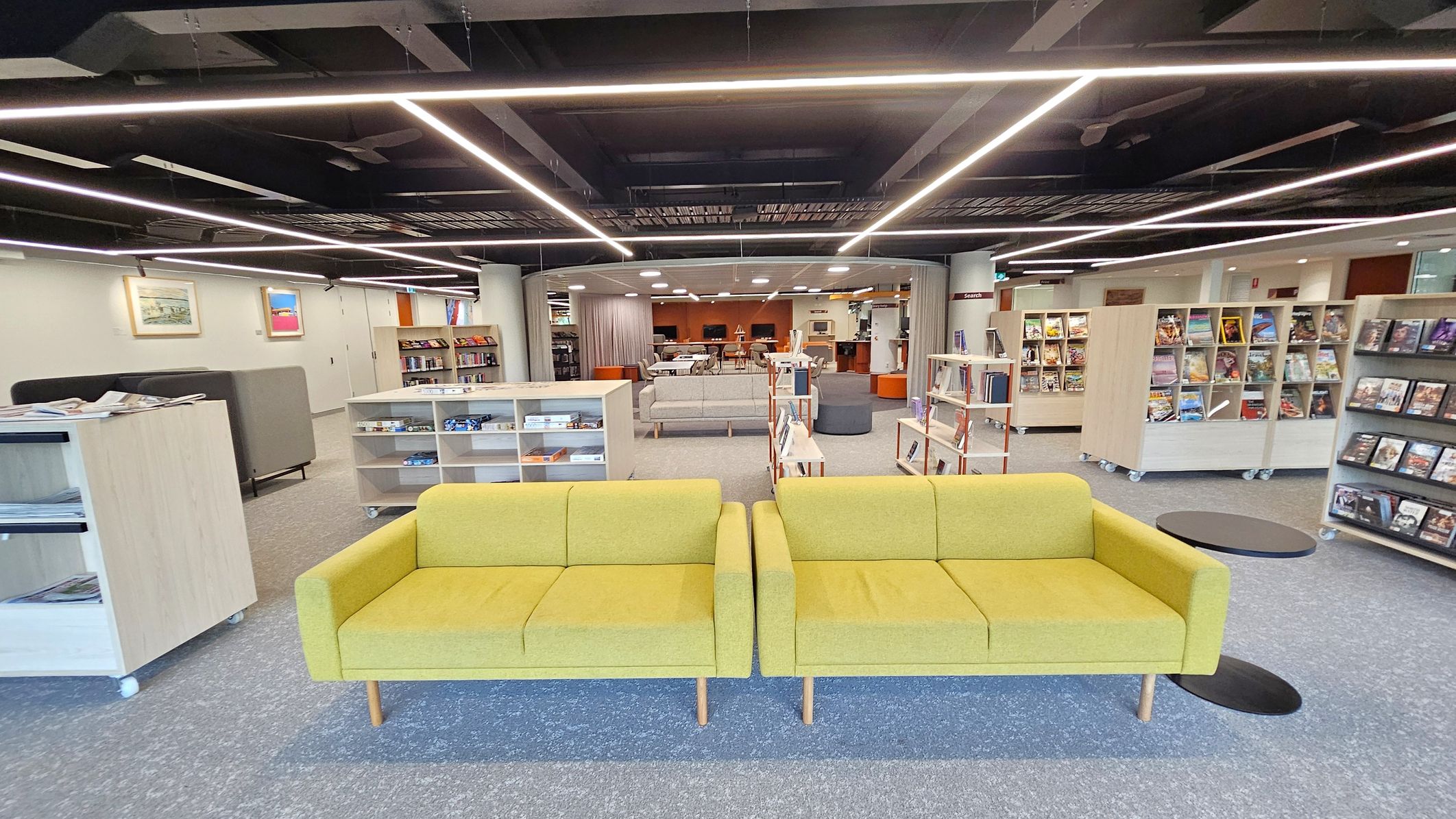 A modern library interior with two yellow sofas in the foreground, placed on a grey carpet. Behind them are low bookshelves filled with books and magazines. The background features more bookshelves, a seating area with grey sofas, and a section with tables and chairs. The ceiling has exposed beams and linear lighting. Walls are adorned with framed artwork, and the floor is carpeted in grey.