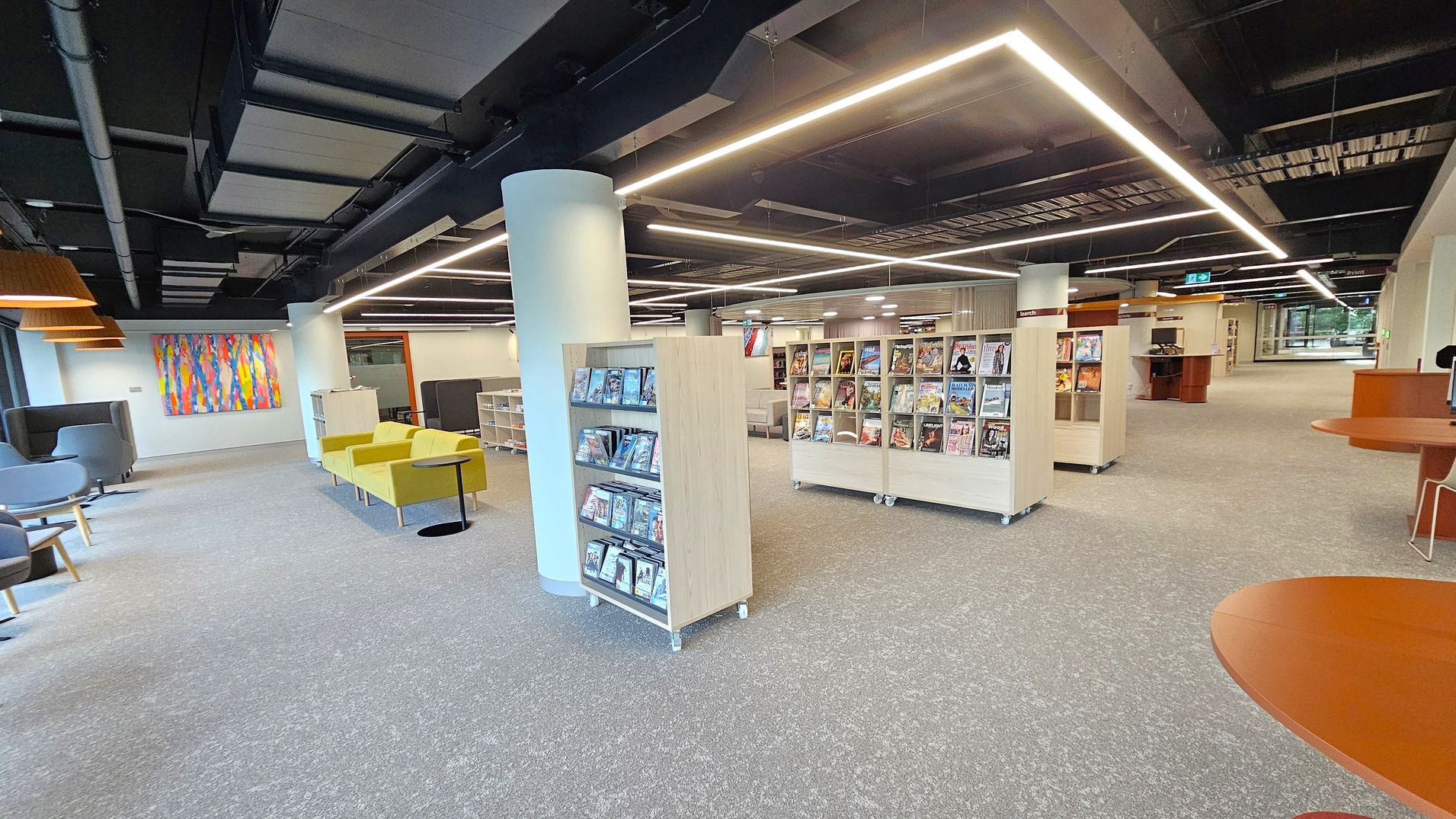A modern library interior with grey carpeted flooring. In the centre, there are shelves displaying magazines and DVDs. To the left, there are grey and yellow seating areas with a colourful abstract painting on the wall. The ceiling features exposed ductwork and linear lighting. On the right, there are more shelves and a wooden table with chairs. The background shows additional seating and a large window.