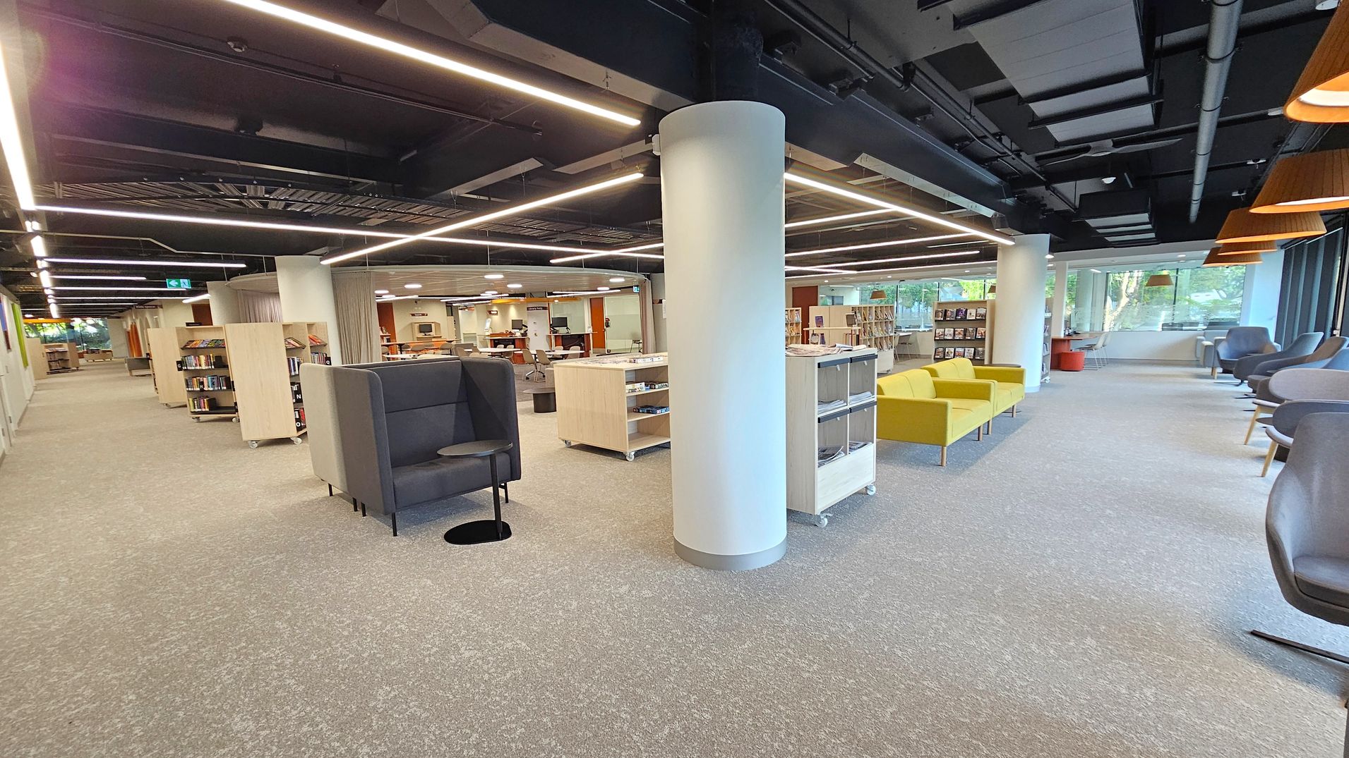 A modern library interior with a grey carpeted floor. On the left, there are bookshelves and a grey high-backed chair with a small round table. In the centre, a white cylindrical column stands amidst more bookshelves and a yellow sofa. The right side features a row of grey armchairs near large windows, allowing natural light in. The ceiling is black with exposed ductwork and linear lighting fixtures.