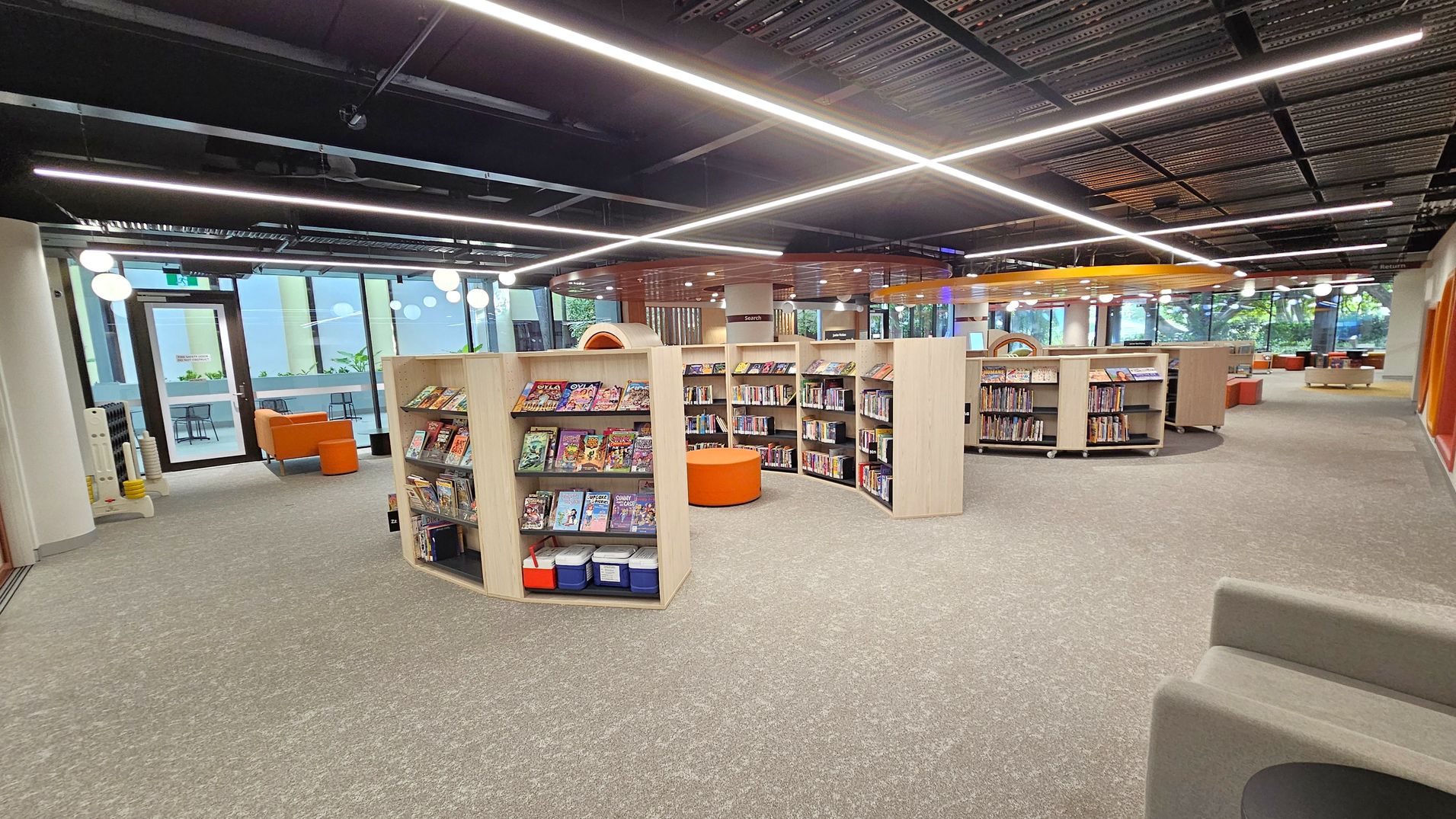 A modern library interior with light wood bookshelves arranged in a semi-circular pattern, filled with books and comics. In the centre, there is an orange circular seating area. The floor is carpeted in grey. Large windows on the left and background allow natural light in, with a view of greenery outside. The ceiling is black with linear lighting fixtures. Orange and grey seating is visible near the windows, and there are round white ceiling lights throughout.