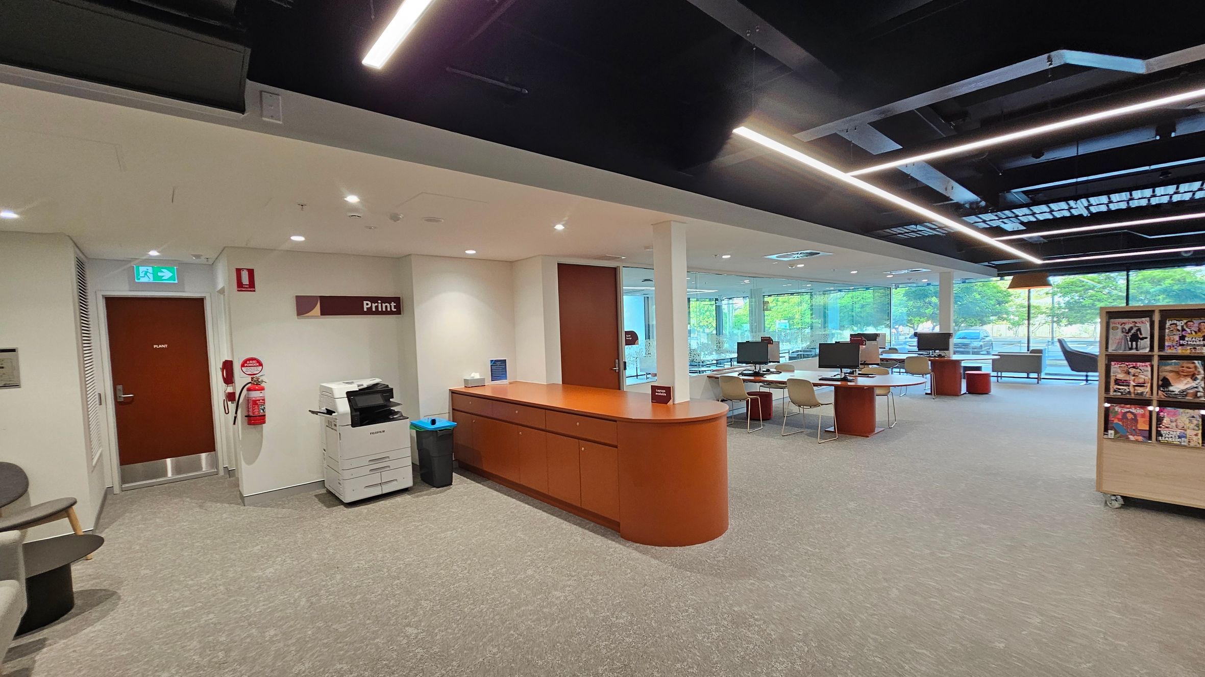 A modern office space with a beige carpeted floor. On the left, there's a wooden counter with a printer and recycling bins nearby. A door marked "Plant" is visible, along with a fire extinguisher. In the centre, there are tables with computers and chairs. The right side features a magazine rack. Large windows in the background allow natural light, and the ceiling has exposed beams with linear lighting.