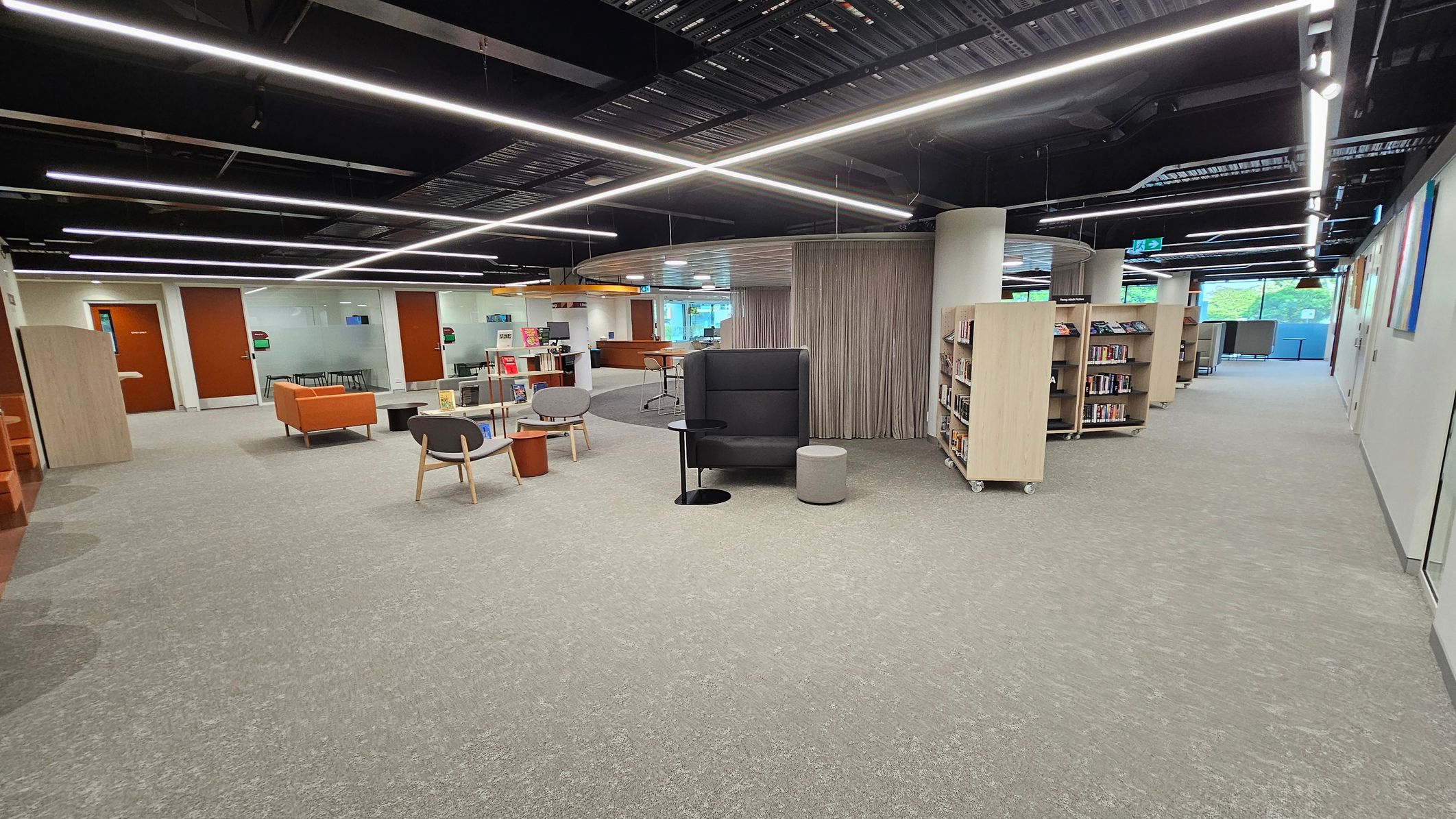 A modern library interior with a light green carpet. On the left, there are orange sofas and grey chairs around small tables. In the centre, a high-backed black chair with a small round table and stool. On the right, bookshelves on wheels line the wall. The ceiling features exposed beams and linear lights. In the background, glass walls and doors lead to additional rooms, with a view of greenery through large windows.