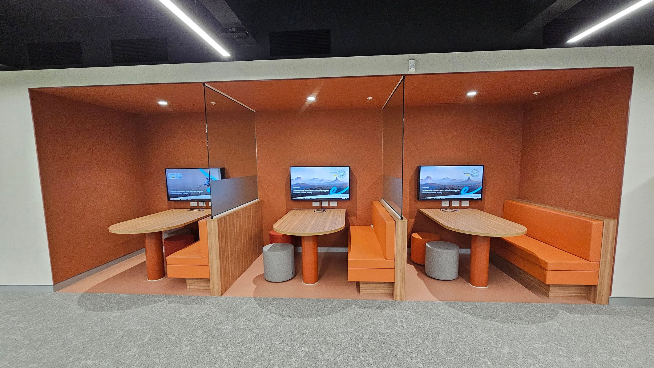 Three adjacent booths with orange walls and ceilings, each containing a wooden table and orange seating. Each booth has a wall-mounted screen displaying a landscape image. The booths are separated by wooden partitions. The floor is grey carpet in front of the booths and orange inside. The ceiling is dark with linear lights above.