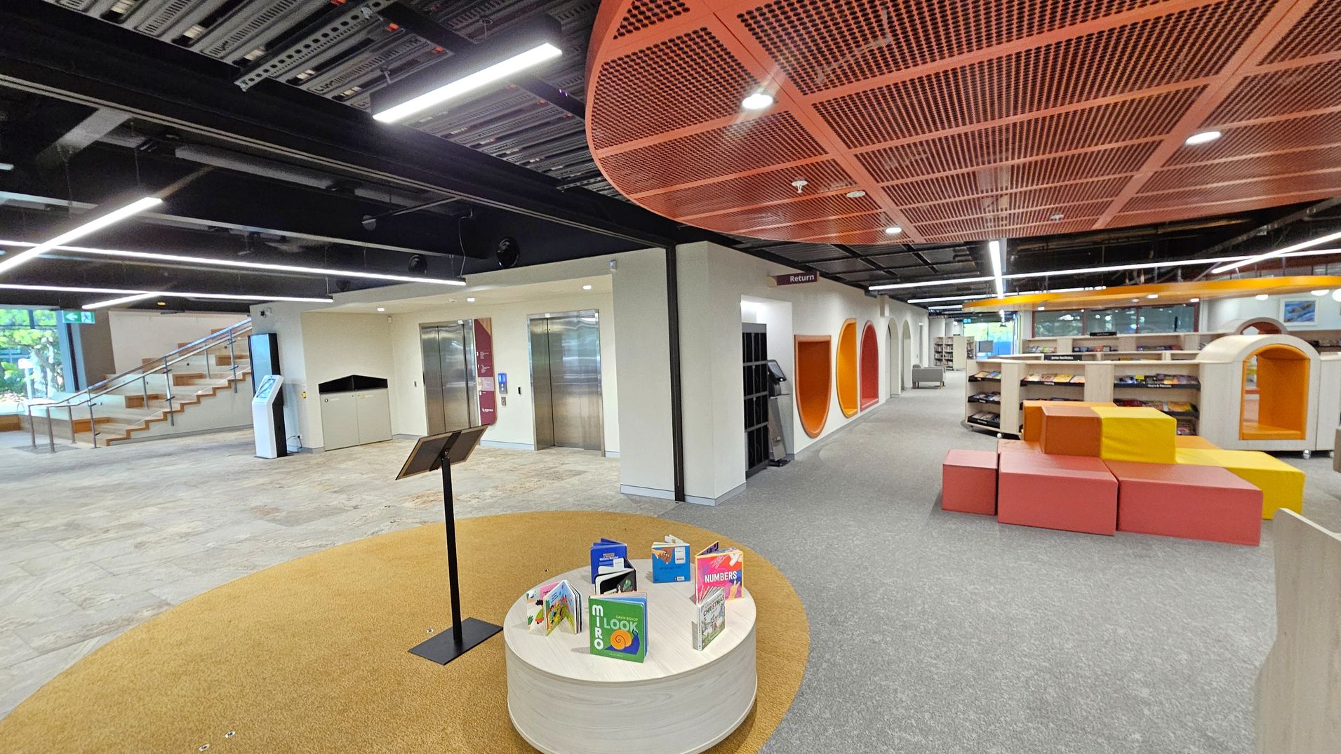 A modern library interior with a grey carpeted floor and an exposed black ceiling with linear lights. On the left, there is a staircase with wooden steps and a digital kiosk. In the centre, a round table displays children's books. On the right, colourful geometric seating blocks in orange and yellow are near bookshelves. The ceiling above has an orange mesh design, and there are circular wall cutouts in orange and red hues.