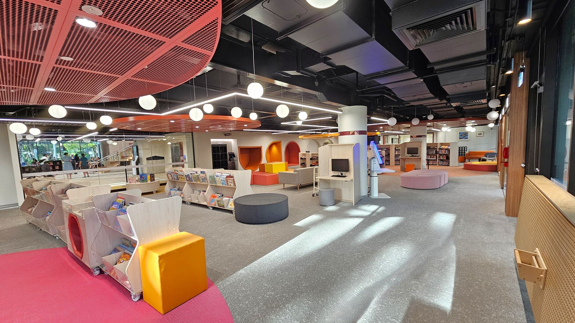 A modern library interior with a colourful and open design. On the left, there are low bookshelves filled with children's books, and a bright orange cube seat on a pink carpet. In the centre, grey and pink circular seating is scattered on a grey carpet. The ceiling features exposed ductwork and hanging spherical lights. On the right, a wooden wall with large windows allows natural light in. The background shows more seating and bookshelves with a variety of books.