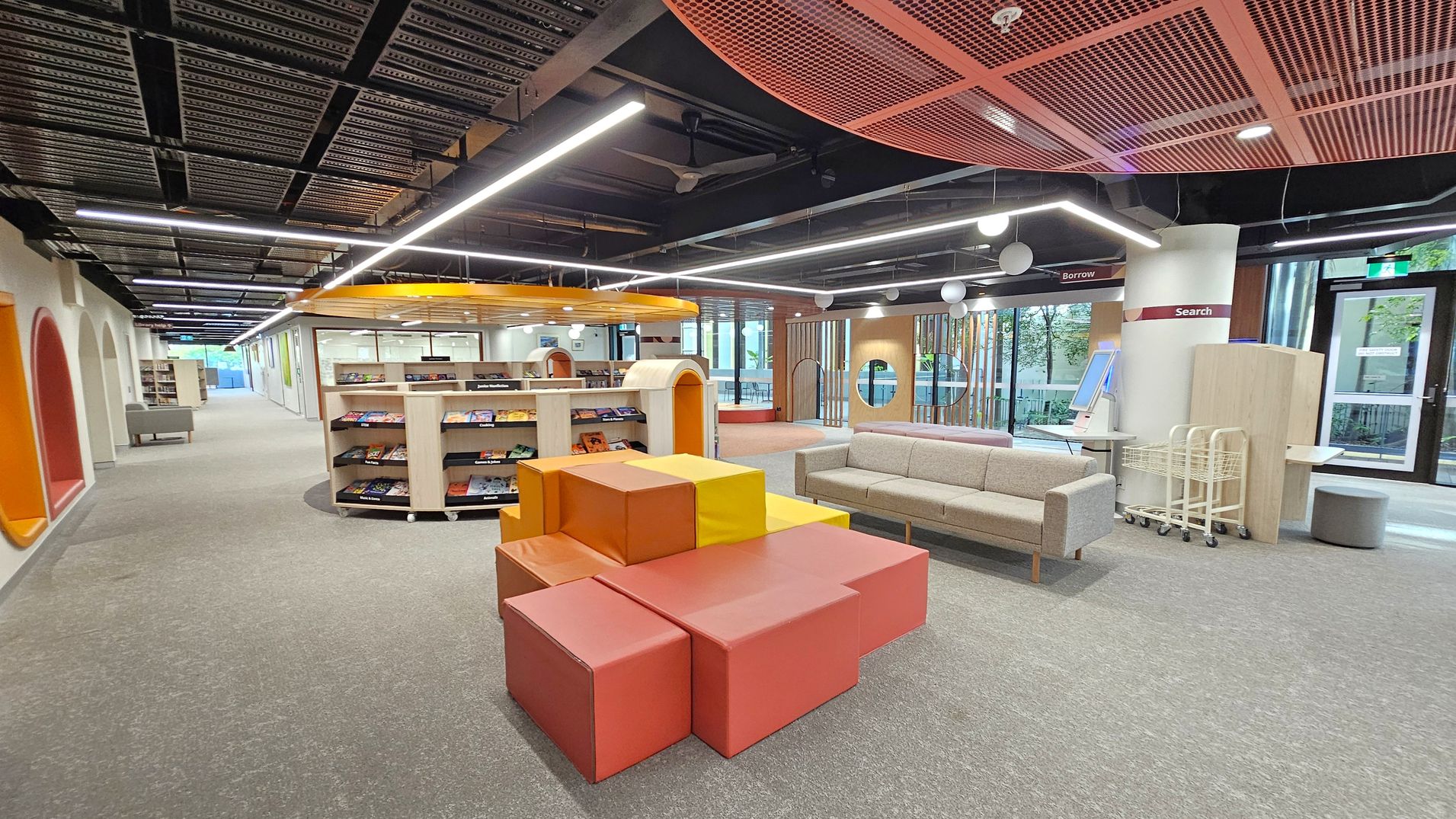 A modern library interior with a grey carpeted floor. In the centre, there are colourful, geometric seating blocks in shades of orange and yellow. To the right, a beige sofa is placed near a column with "Search" and "Borrow" signs. Bookshelves with magazines are visible in the middle. The ceiling features exposed black beams and linear lighting. Large windows on the right allow natural light, and there are circular cutouts in the wooden wall panels.