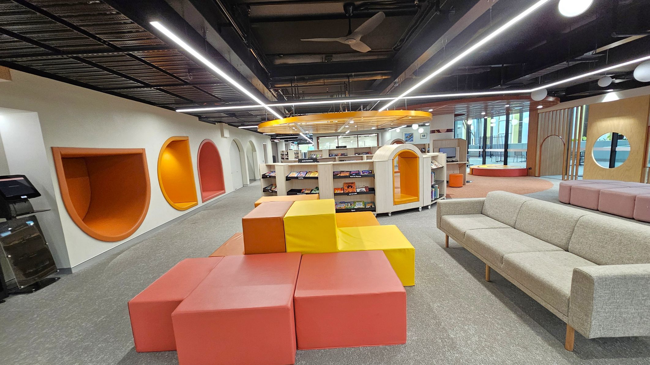 A modern library interior with a grey carpeted floor. On the left, there are colourful, semi-circular reading nooks in orange and red. In the centre, there are orange and yellow cube seats arranged in a stepped formation. Bookshelves with books are visible behind them. On the right, a light grey sofa is placed near a circular window. The ceiling is black with exposed ductwork and linear lighting. The background features large windows and additional seating areas.