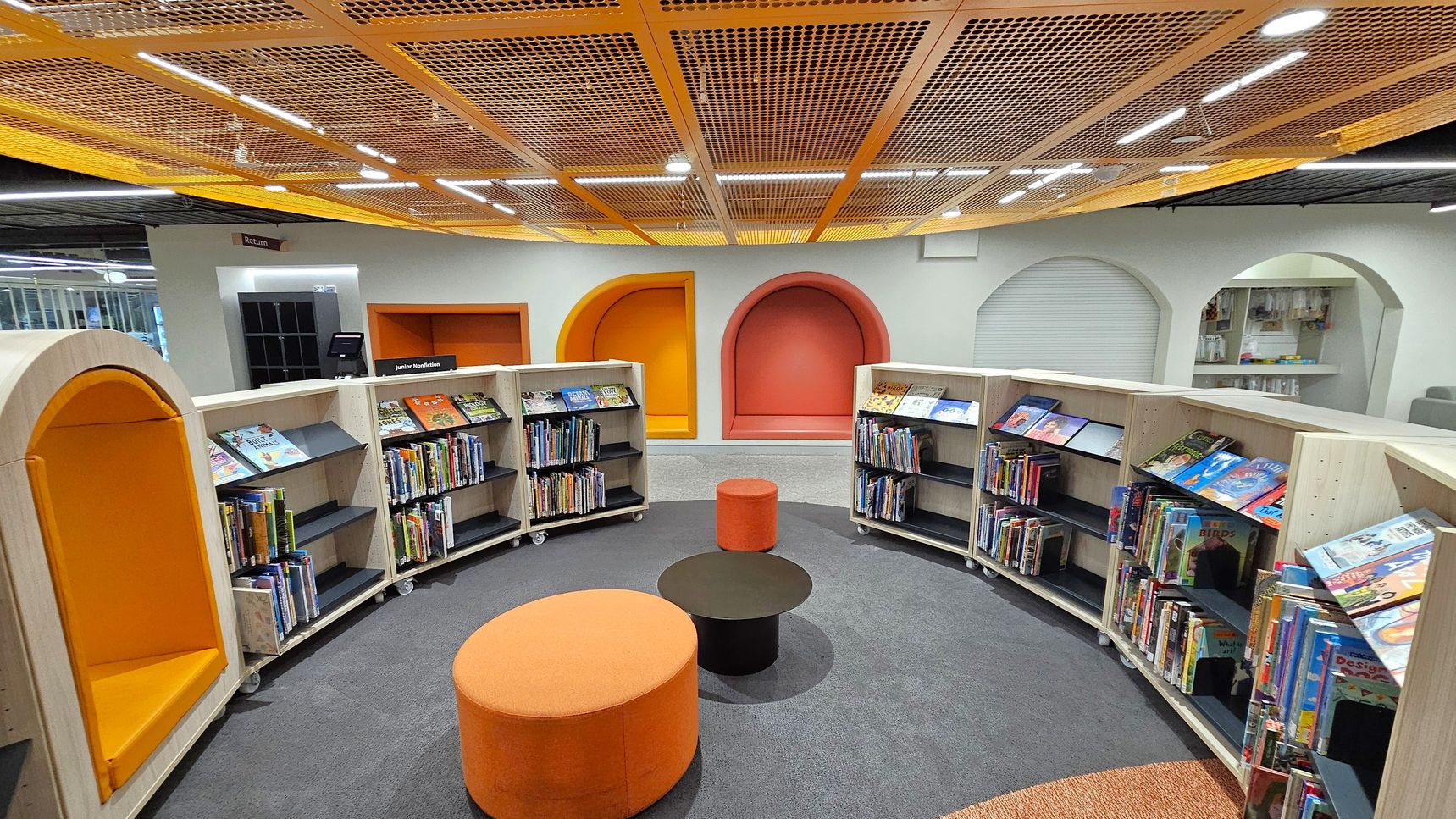 A modern library space with curved bookshelves filled with children's books. In the centre, there are round orange and black stools on a grey carpet. The back wall features colourful, arched alcoves in orange and red. The ceiling has a grid pattern with orange accents and integrated lighting. A sign for "Junior Nonfiction" is visible, and there's a book return area on the left.