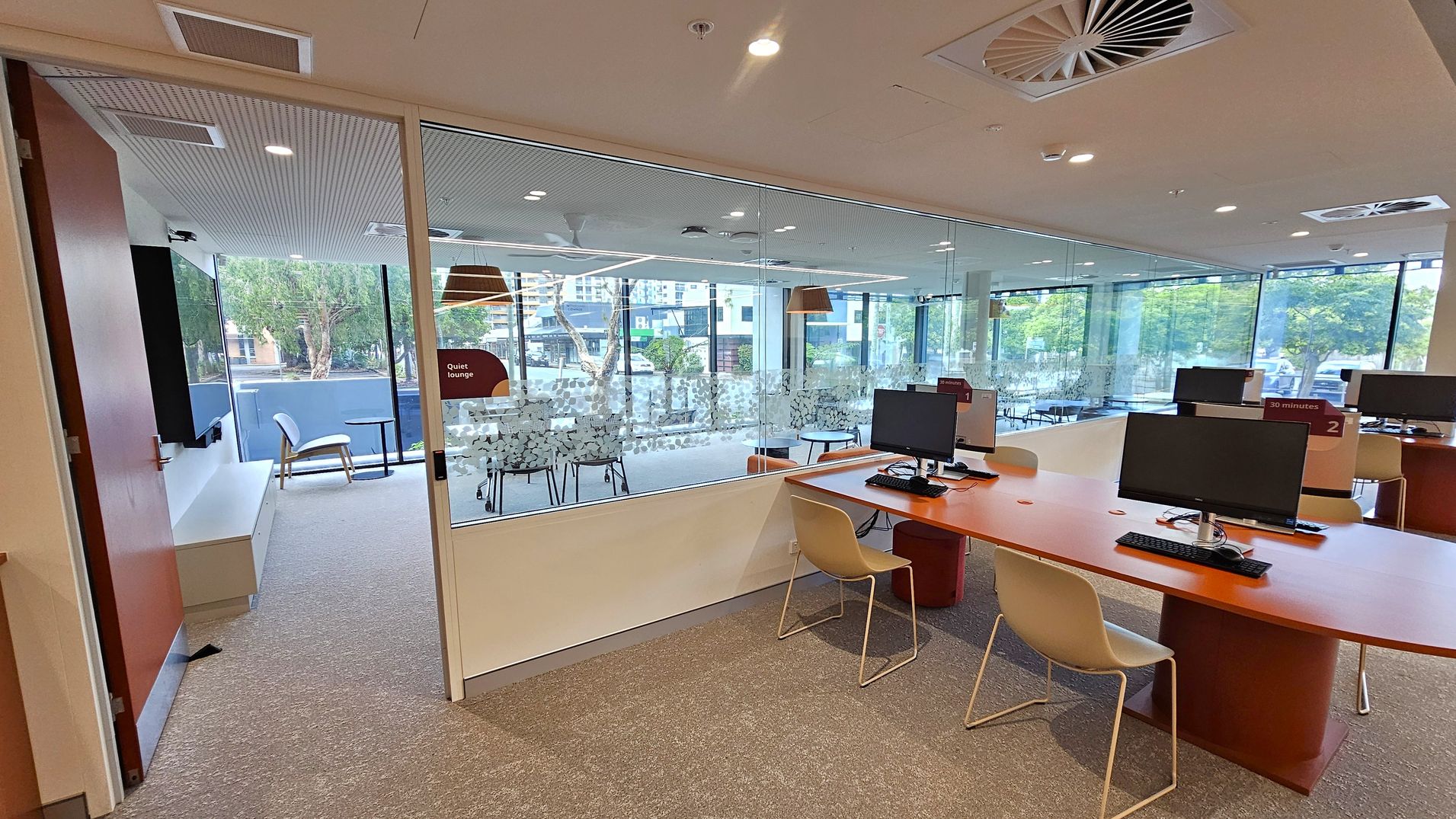 A modern office space with a glass-walled room labelled "Quiet lounge" on the left, containing a chair and a small table. In the foreground, there are several computer workstations with monitors and keyboards on wooden desks, each accompanied by a chair. The floor is carpeted, and the ceiling features recessed lighting and air vents. Large windows in the background provide a view of trees and buildings outside.