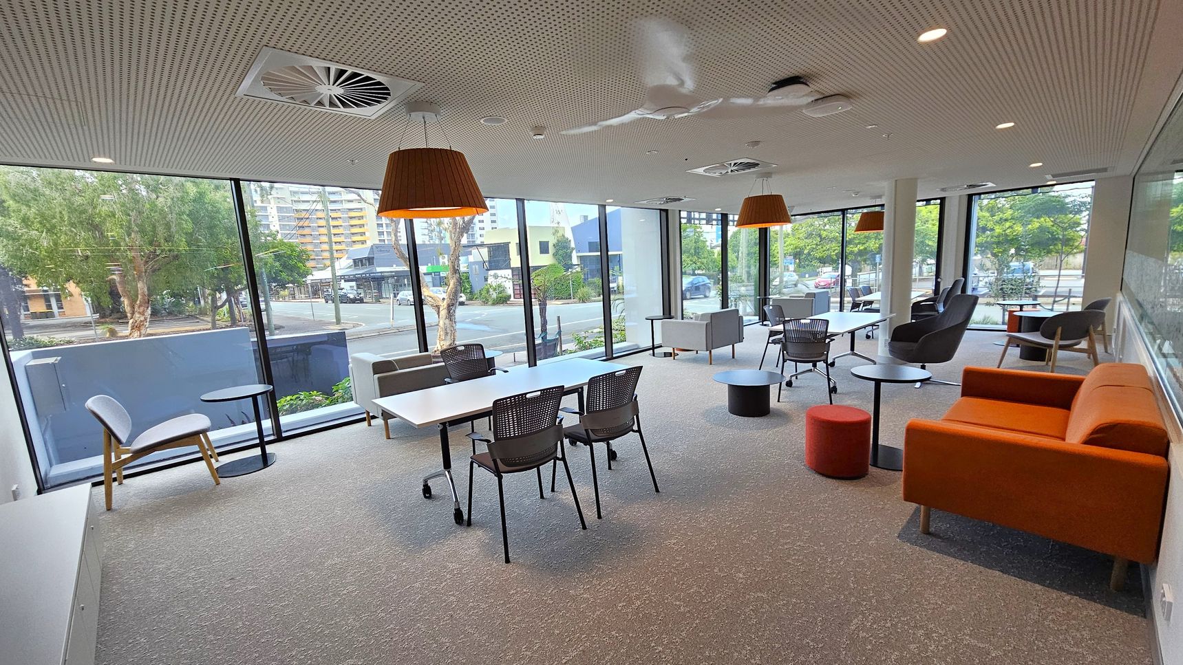 A modern office lounge with large floor-to-ceiling windows on the left and right, offering views of trees and buildings outside. The room features a mix of seating, including grey chairs, black mesh chairs, and an orange sofa. White tables are arranged in the centre, with small round tables scattered around. The ceiling has recessed lighting, air vents, and large pendant lights. The floor is carpeted in grey.