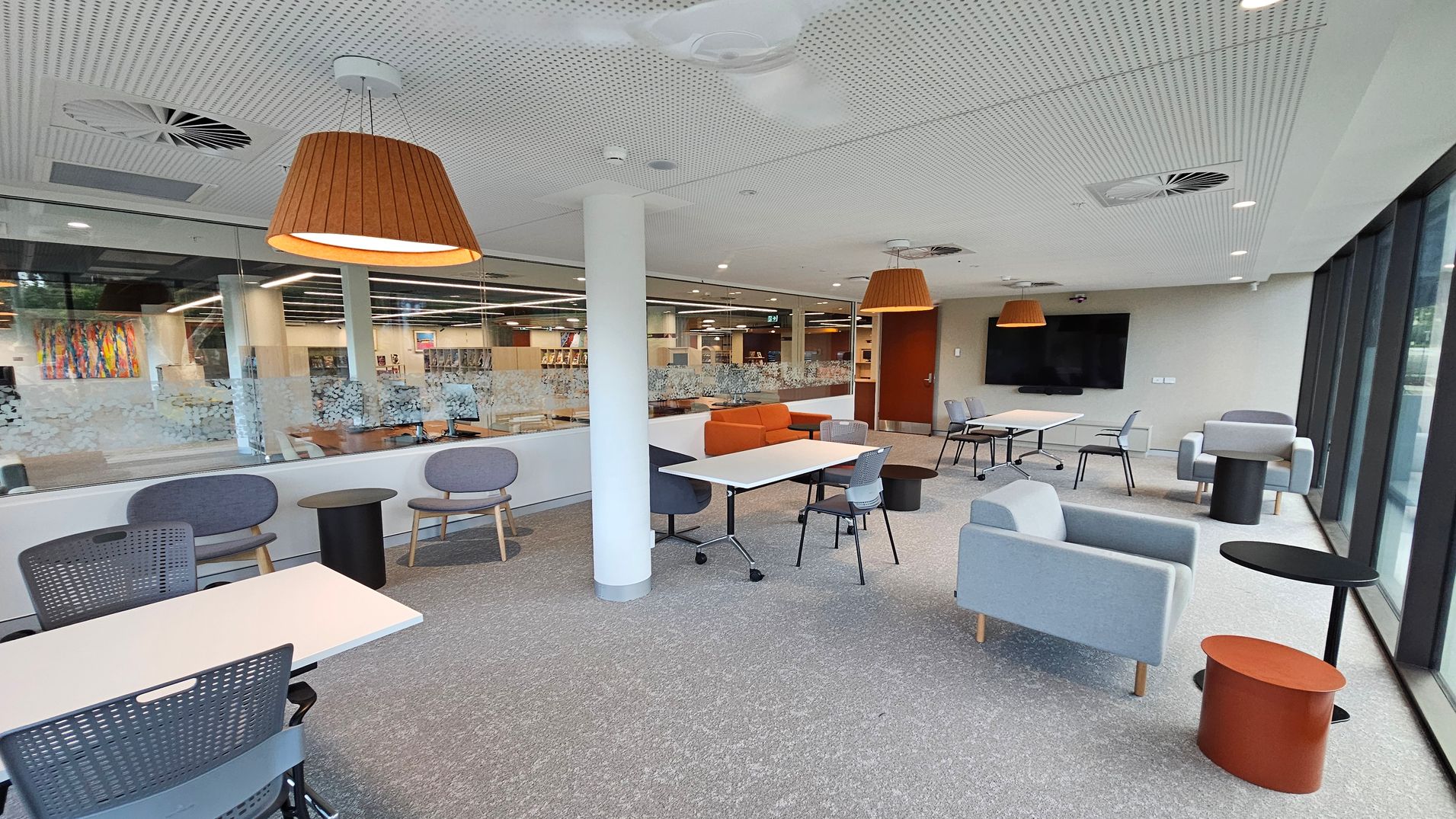 A modern, open-plan office space with grey carpeted flooring. On the left, there are round tables with grey chairs and a glass wall with a view into another room. In the centre, a white column supports the ceiling, which has recessed lighting and wooden pendant lamps. On the right, there are light blue armchairs, small round tables, and a large wall-mounted screen. The far wall features large windows allowing natural light to enter.