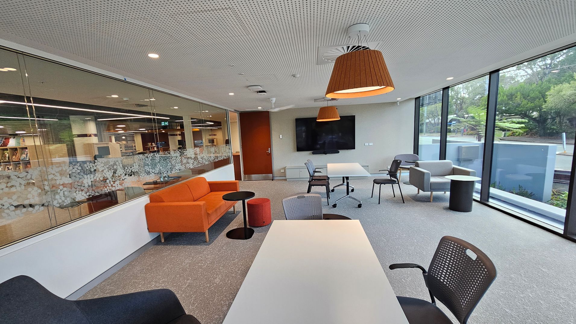 A modern office space with a perforated white ceiling and grey carpeted floor. On the left, there's a glass wall with a decorative pattern, revealing a library area. An orange sofa and small round tables are nearby. In the centre, a large screen is mounted on the wall with a table and chairs in front. On the right, large windows offer a view of greenery outside. Pendant lights hang from the ceiling, and a grey armchair with a side table is positioned near the windows.